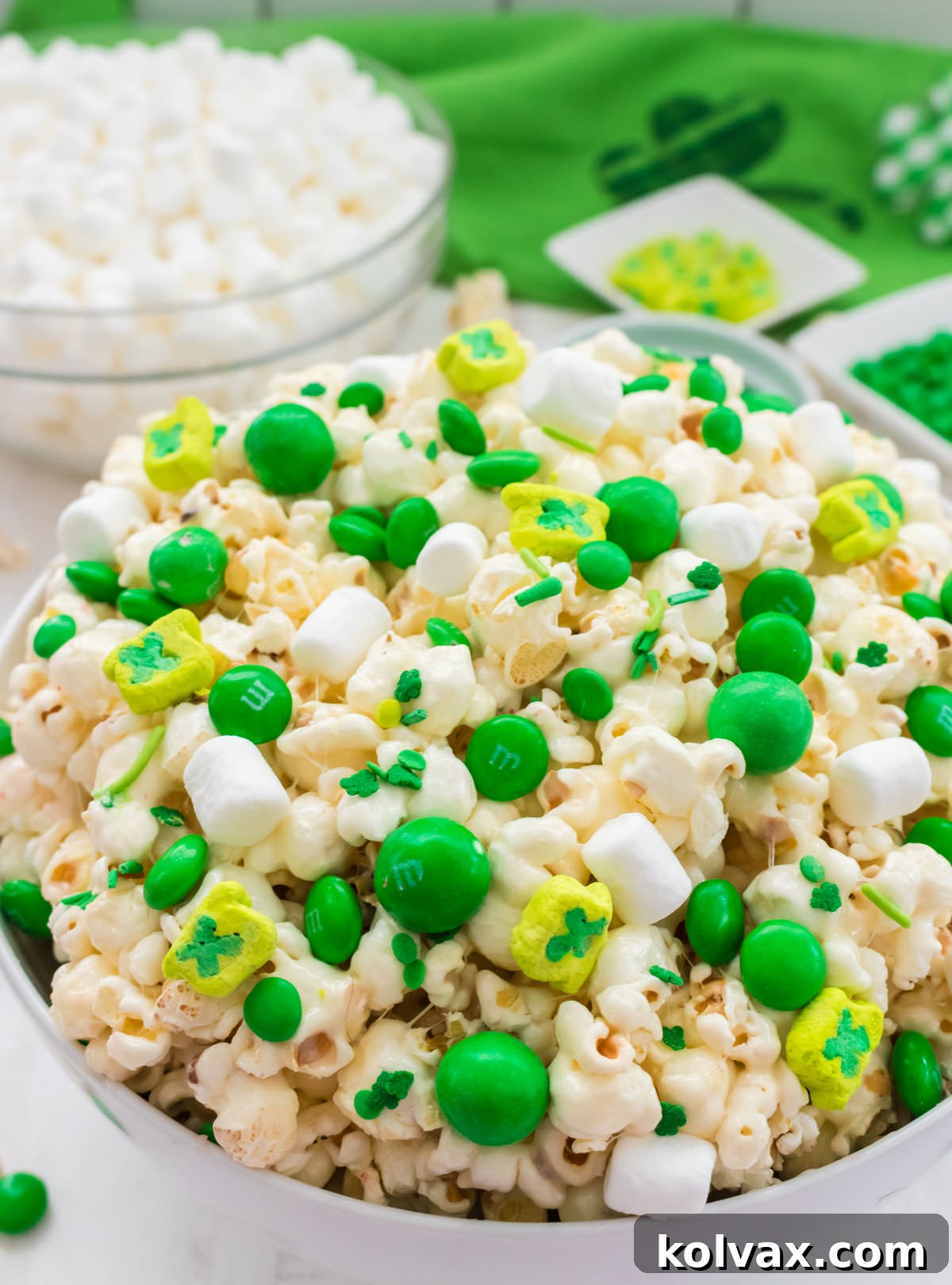 Close up on a beautiful white serving bowl filled to the brim with festive St. Patrick's Day Popcorn, featuring green M&M's and shamrock marshmallows. In the blurred background, a clear bowl holds extra mini marshmallows and a festive St. Patrick's Day themed table linen adds to the holiday ambiance.