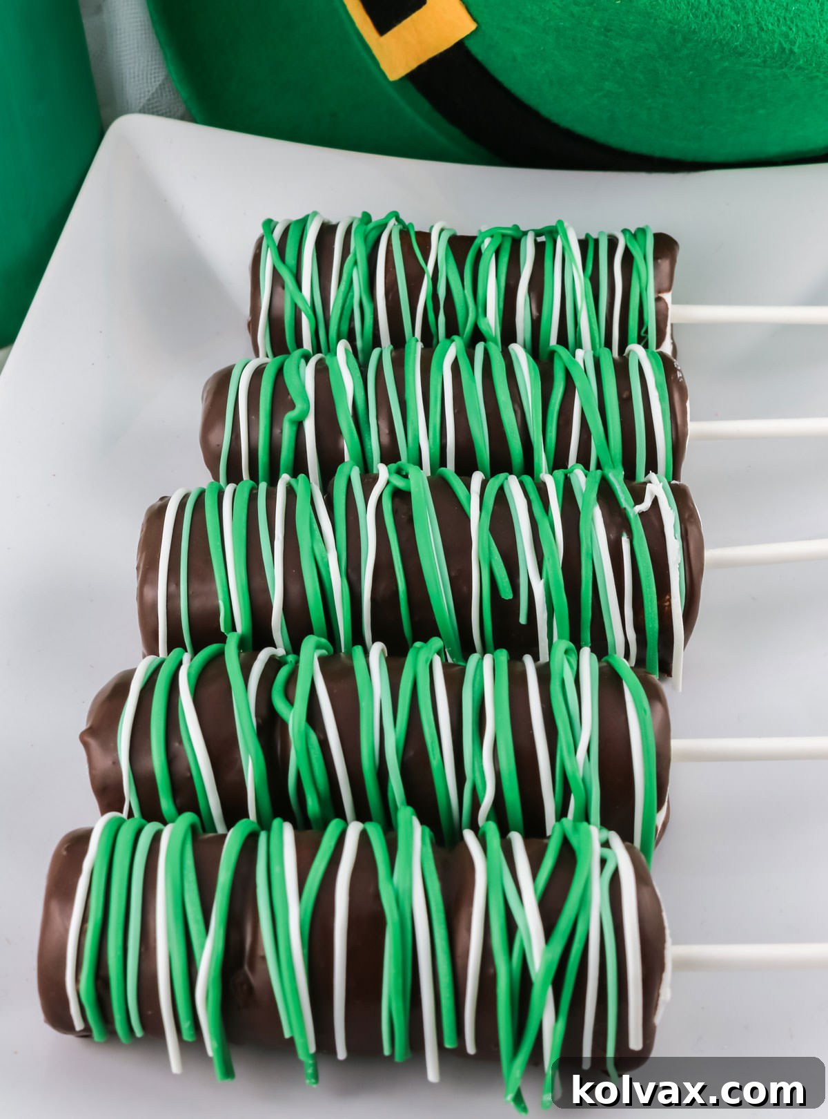 Five St. Patrick's Day Marshmallow Pops laying on a white serving platter in front of a Leprechaun hat.