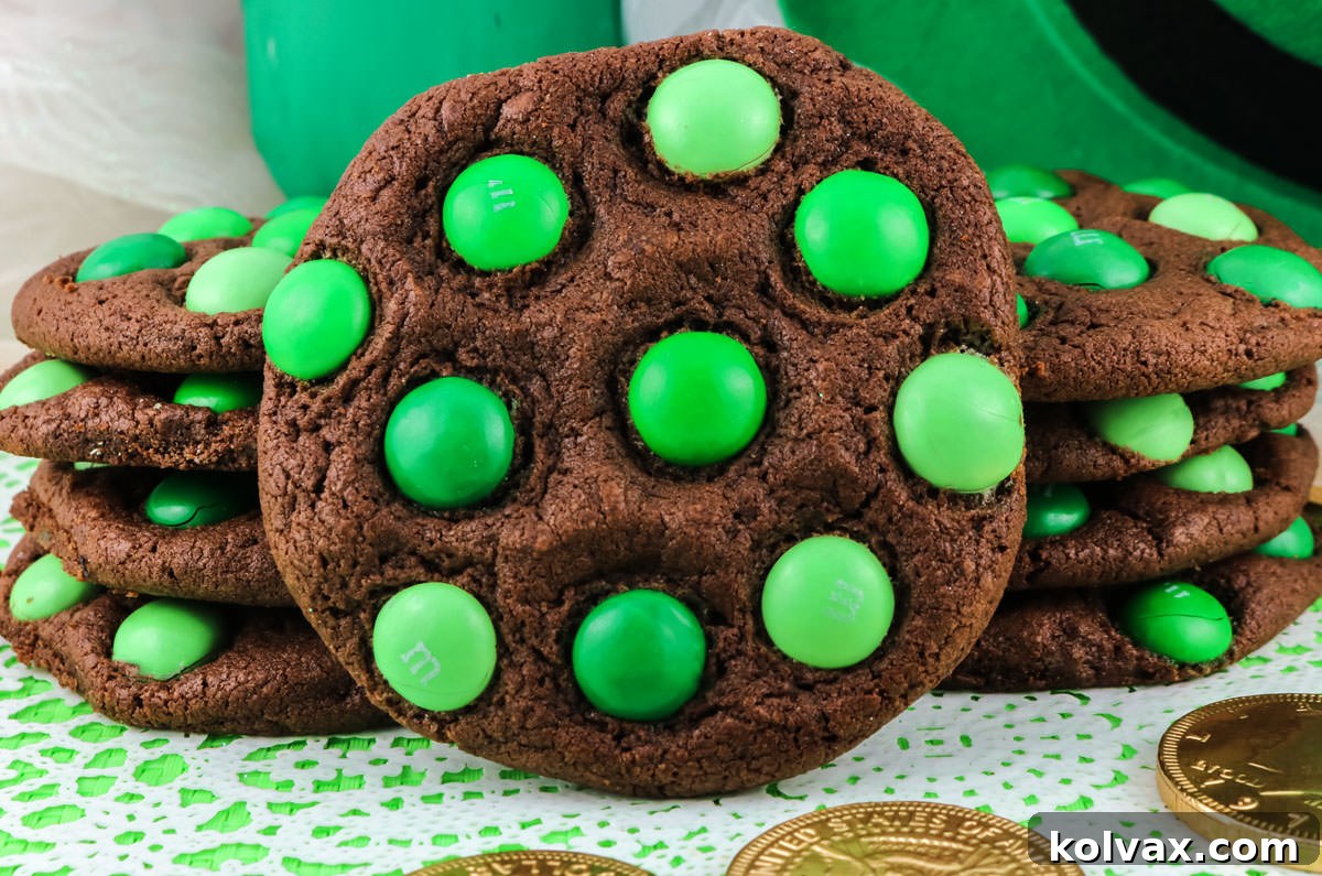 A closeup of a St. Patrick's Day Mint M&M cookie leaning against two other stacks of cookies surrounded by gold coins and a leprechaun hat. The cookies are dark chocolate with green mint M&M's.