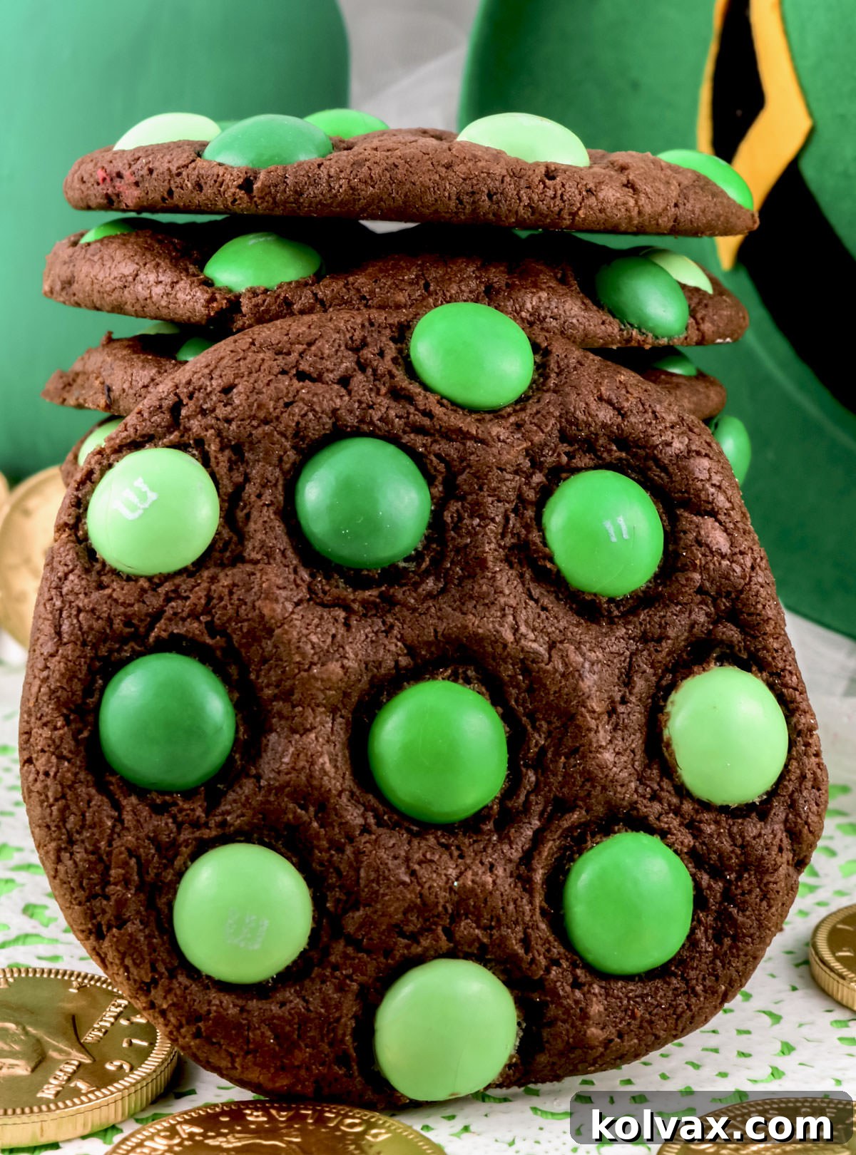 Closeup on a stack of St. Patrick's Day Mint M&M Cookies sitting on a white piece of lace surrounded by gold pieces and in front of a leprechaun hat.