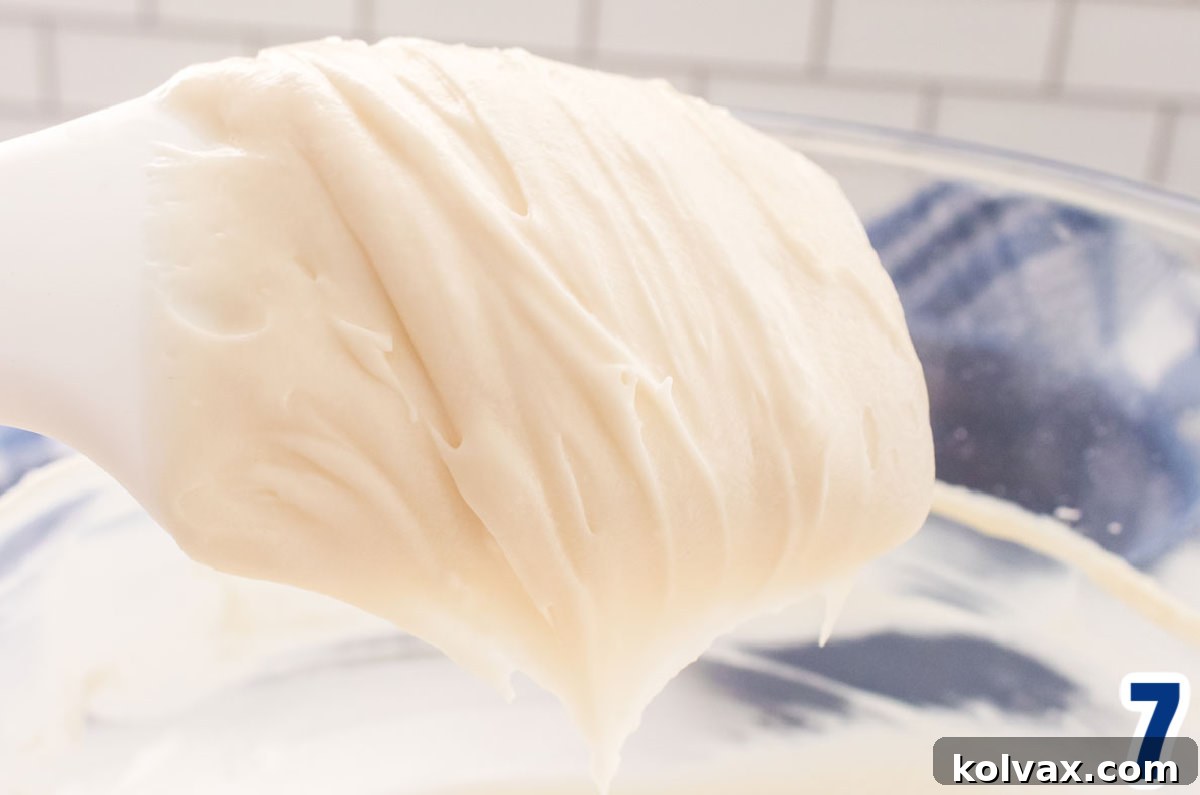 Close up on a white spatula covered with Cream Cheese Icing that is hovering above a glass mixing bowl that is sitting on a white table.