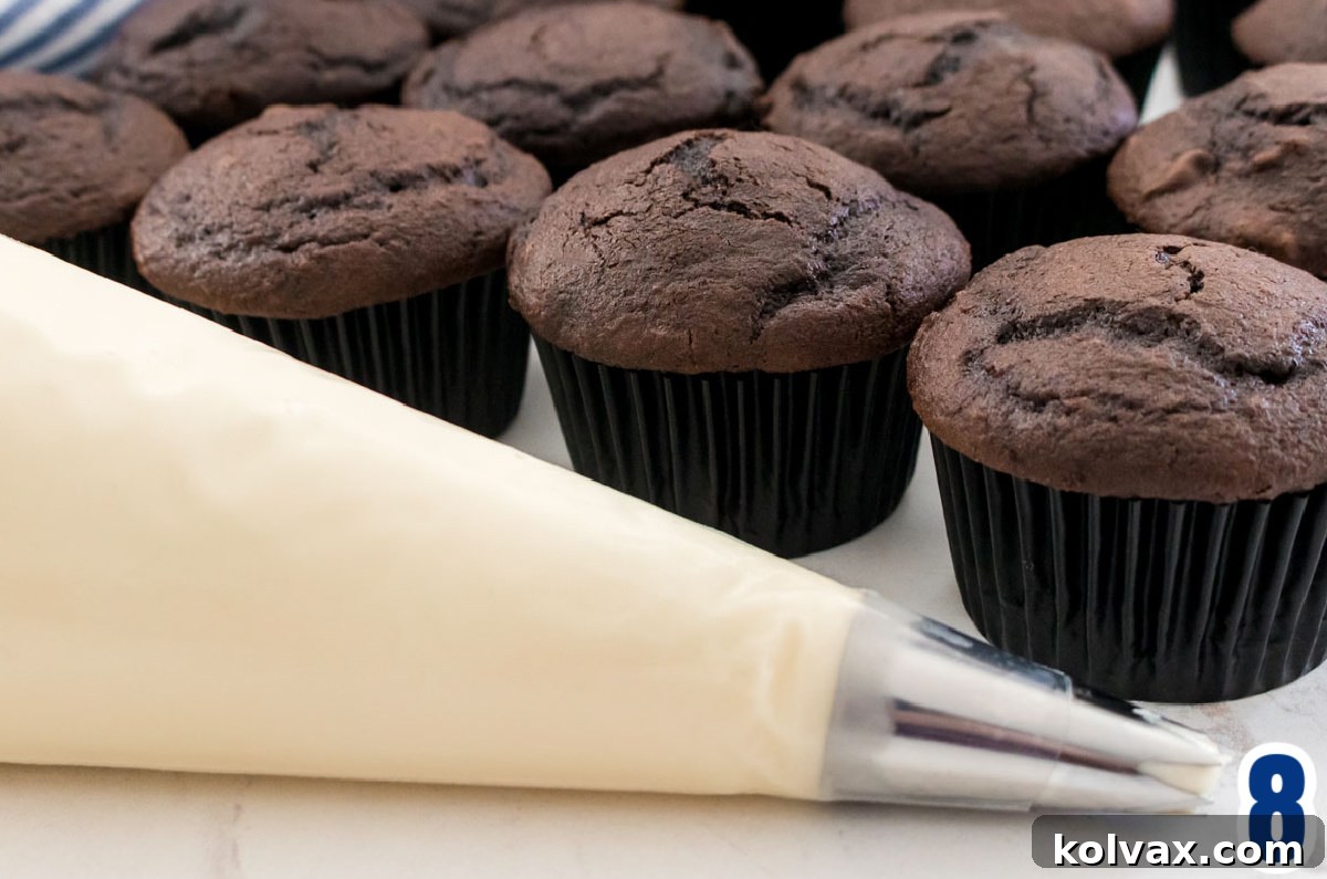 A frosting decorating bag filled with Cream Cheese Frosting sitting on a white table in front of a batch of unfrosted chocolate cupcakes.