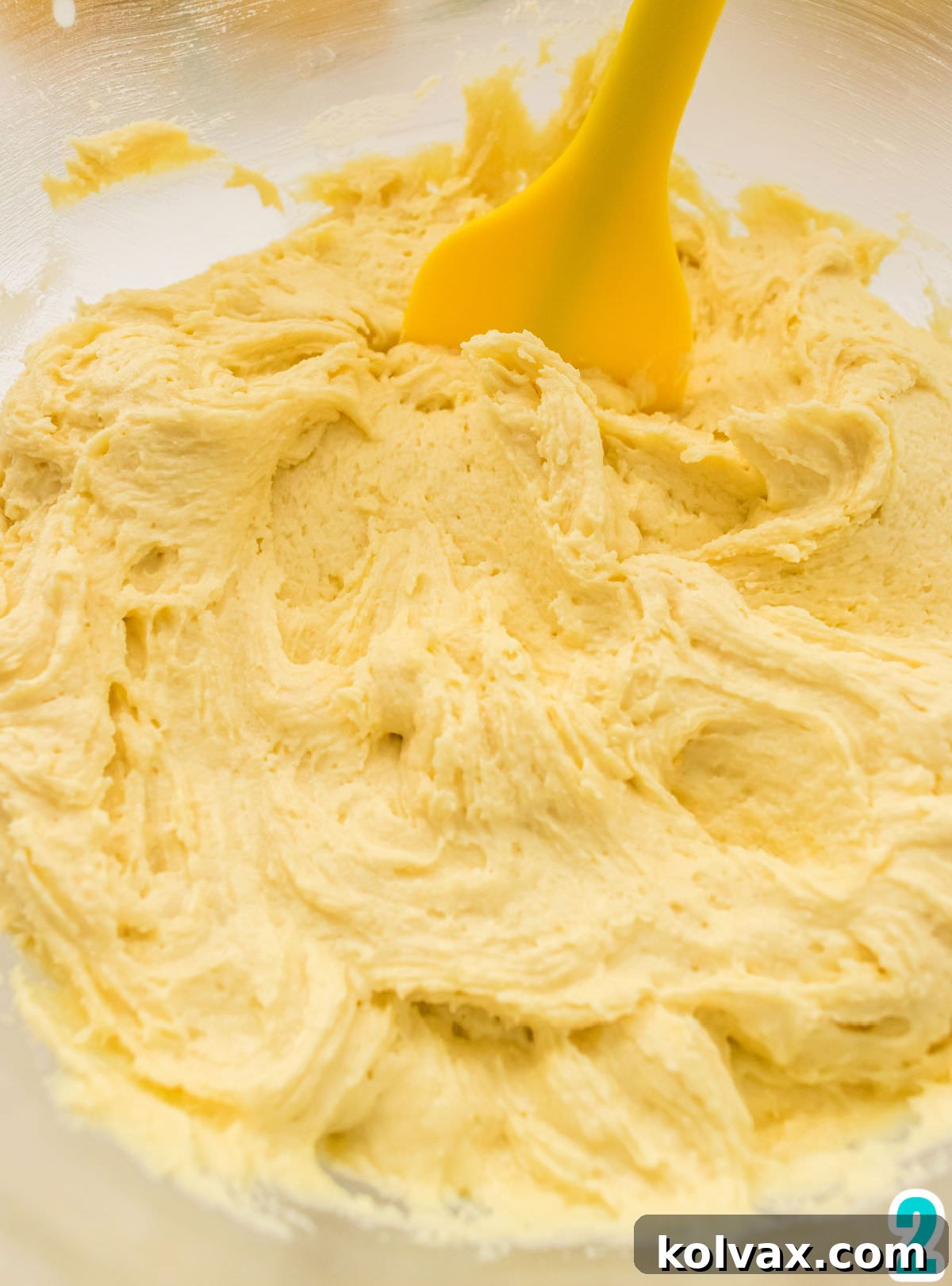 A close-up view of a clear glass mixing bowl filled with creamy, pale yellow lemon cookie dough, with a yellow silicone spatula resting inside. The dough appears smooth and well-mixed, ready for chilling.