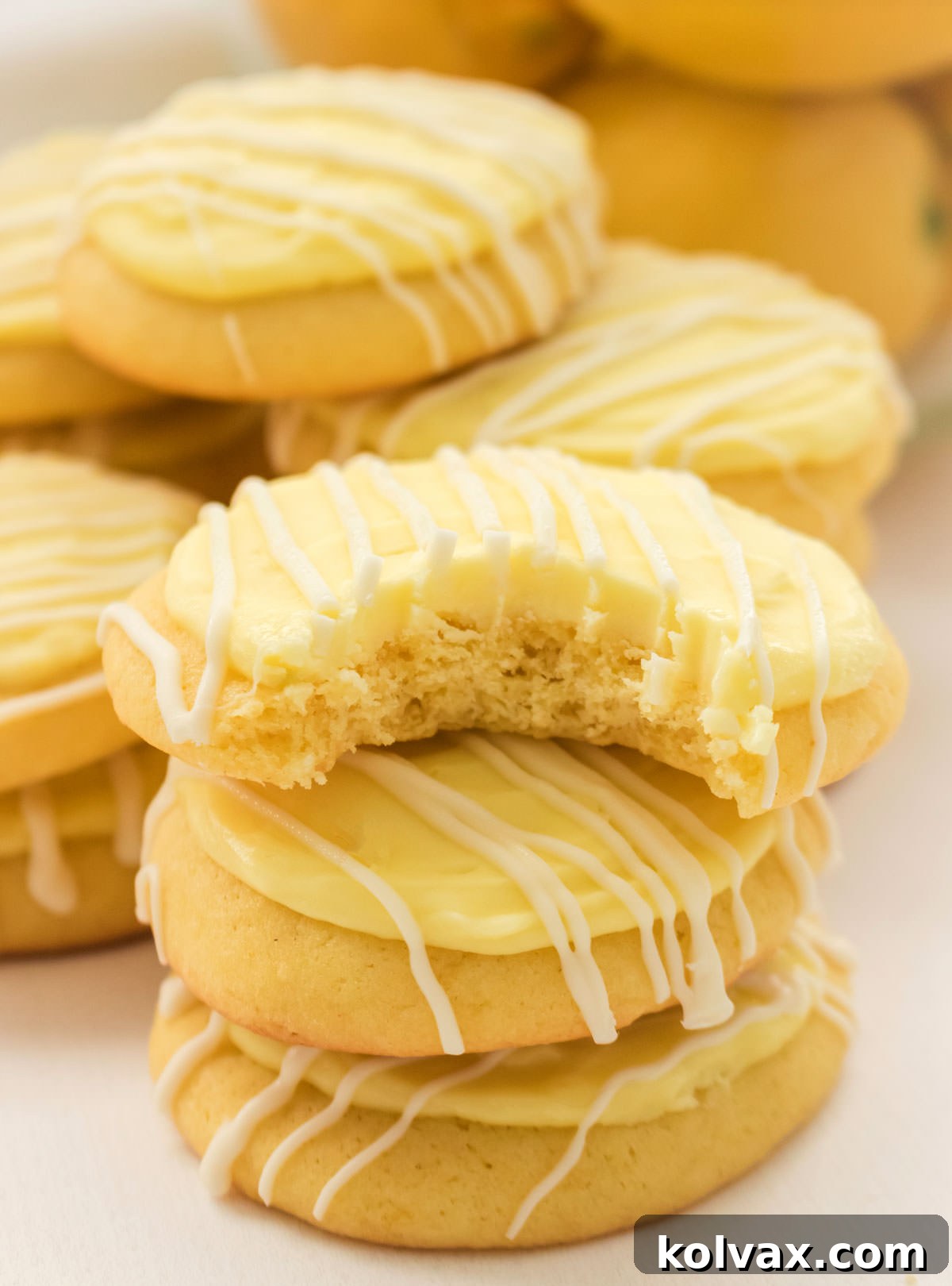 A close-up photograph of a stack of fluffy Lemon Cookies with Lemon Frosting, with the top cookie having a bite taken out, revealing its soft, cakey interior and thick layer of creamy lemon frosting.