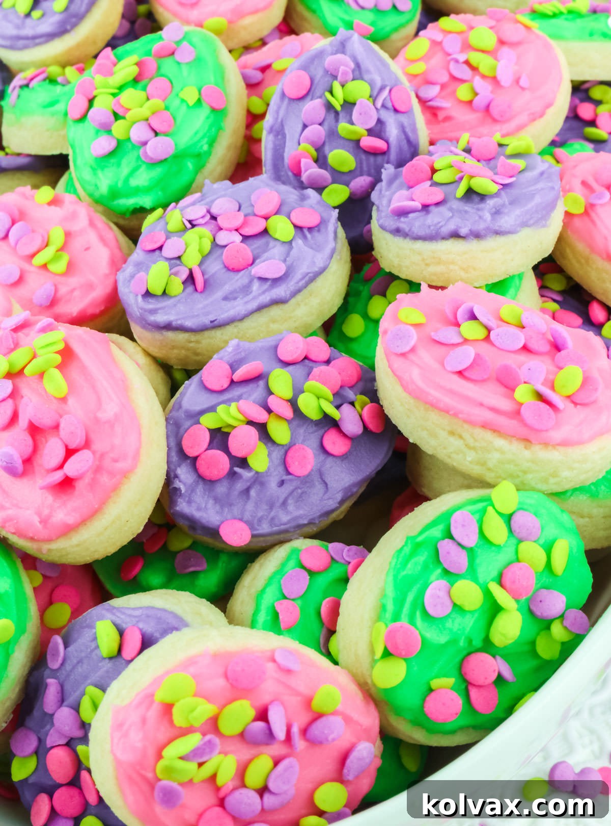 Close up of a white serving bowl generously filled with an assortment of purple, pink, and green Easter Egg Cookies, all adorned with pastel sprinkles.
