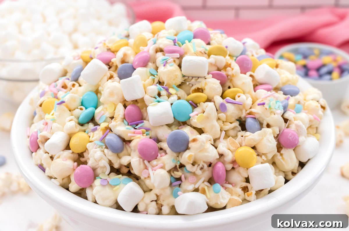 A close-up of Springtime Celebration Popcorn in a white bowl, with a background of marshmallows and Easter M&M's in glass containers.