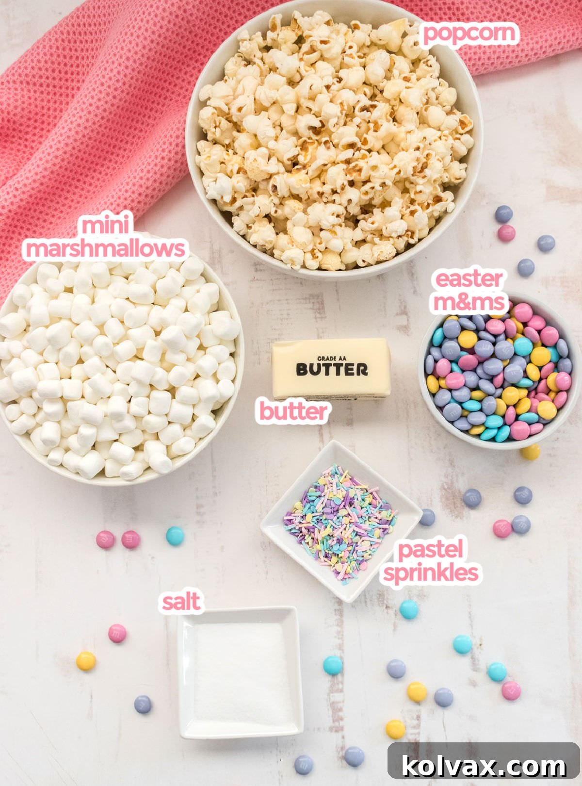 All the essential ingredients for Springtime Celebration Popcorn, including popcorn kernels, butter, mini marshmallows, Easter M&M's, and pastel sprinkles, laid out on a surface.