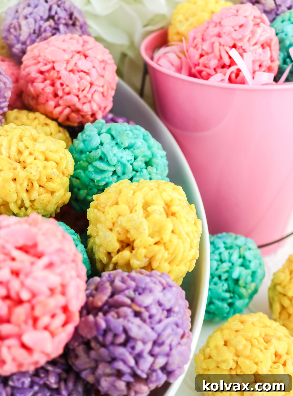 Closeup of a white serving bowl filled with colorful Easter Rice Krispie Treat Bites with a pink pail also filled with Bites sitting right next to it, decorated with spring flowers.