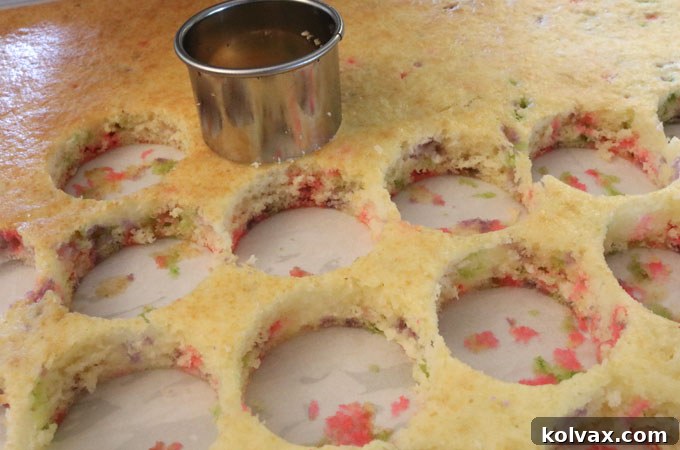 A hand using a round cookie cutter to cut mini cake bites from a cooled confetti cake sheet.