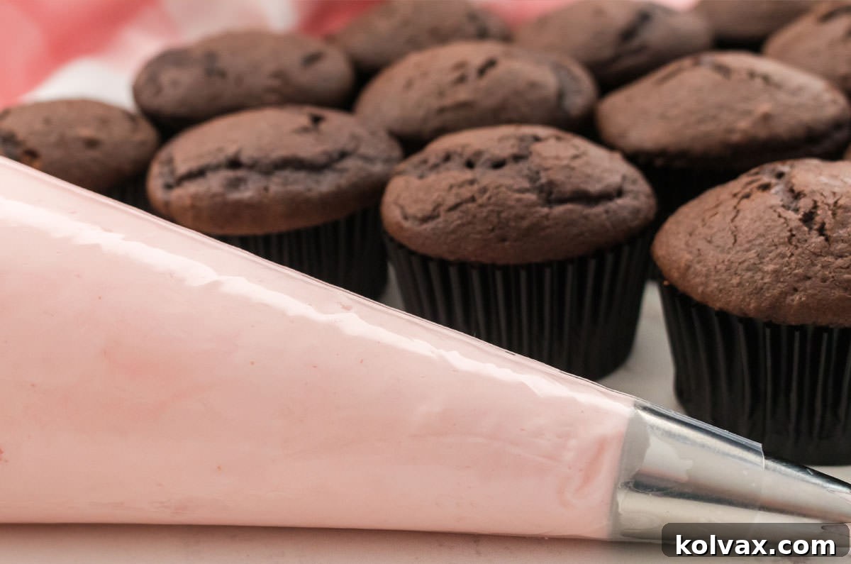 A piping bag filled with vibrant Pink Lemonade Frosting, resting in front of a tray of unfrosted chocolate cupcakes, ready for decoration.
