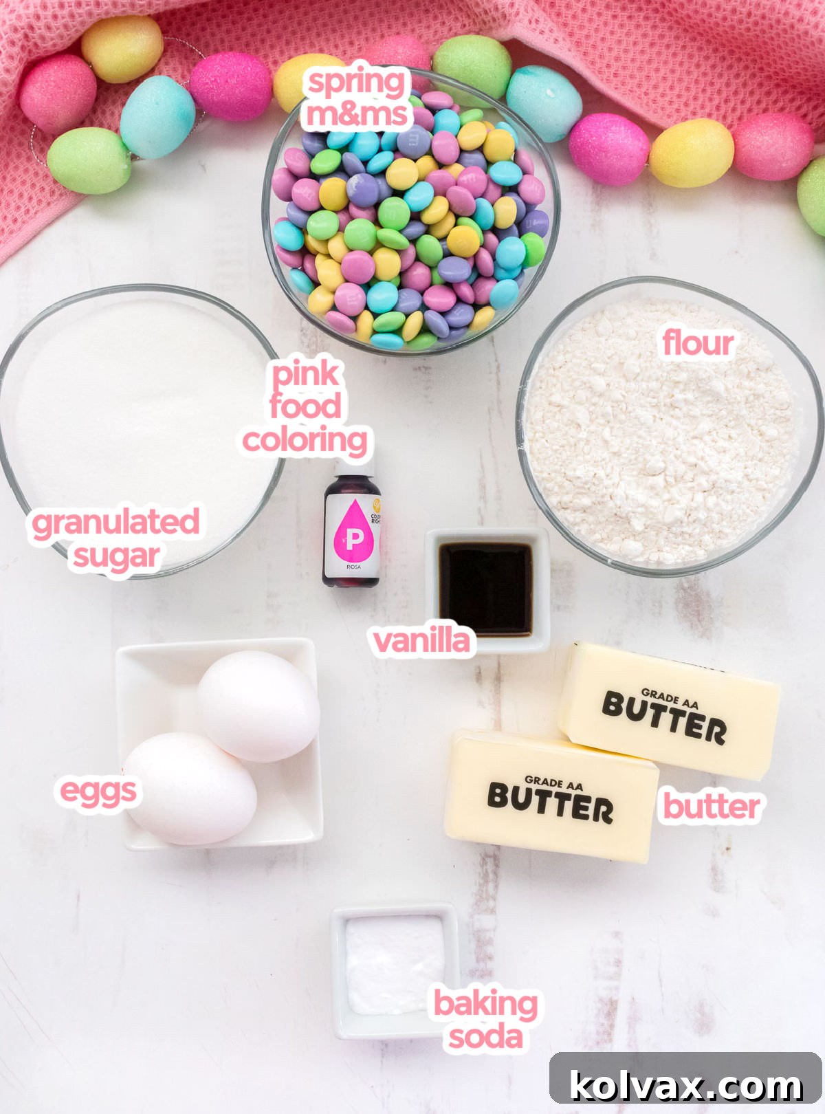 A flat lay of all the essential ingredients needed to make Easter M&M Sugar Cookies, including sticks of butter, granulated sugar, fresh eggs, vanilla extract, baking soda, all-purpose flour, a small bottle of pink food coloring, and a generous bag of Spring M&M's.
