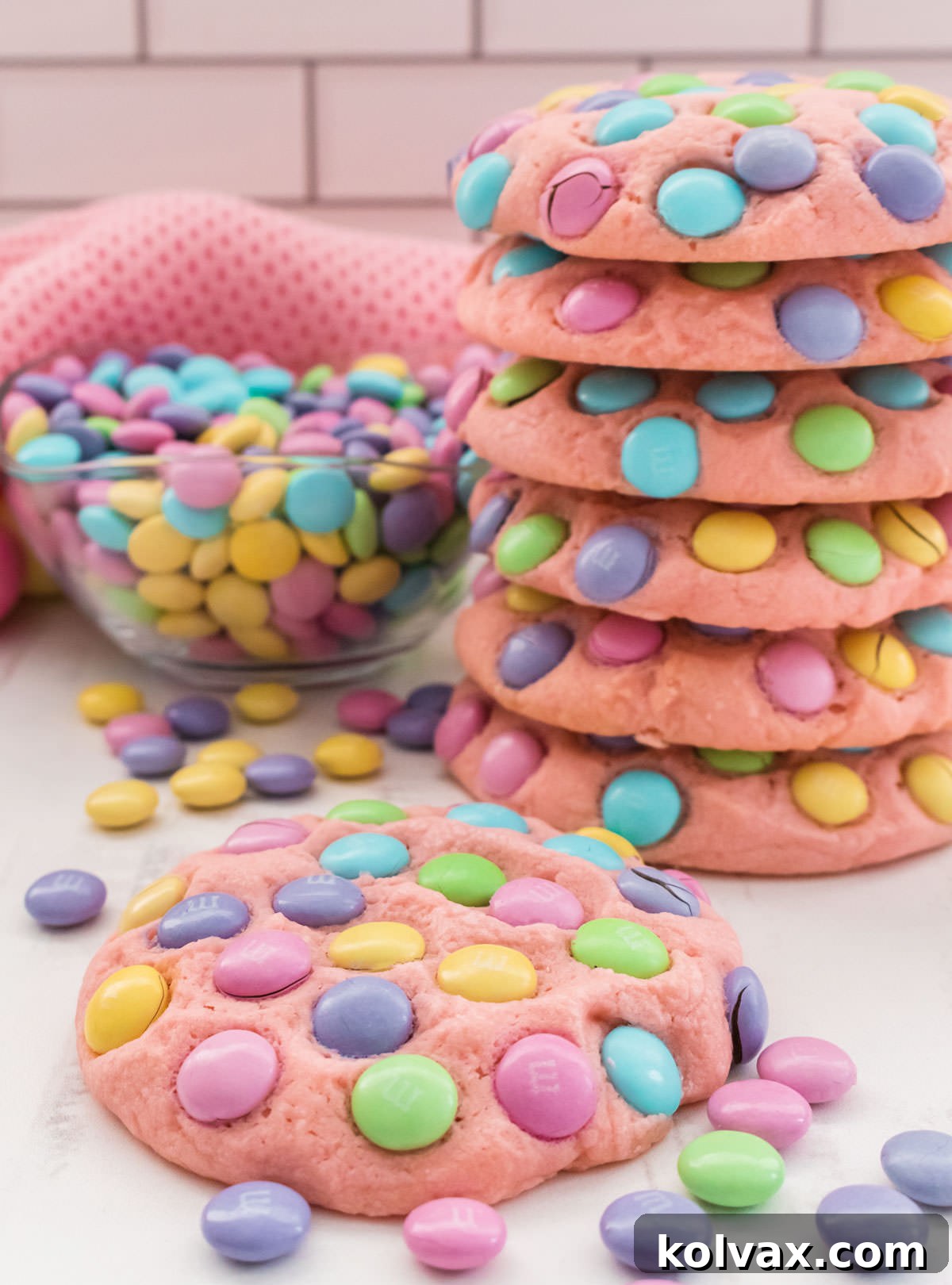 A charming close-up of a single Easter M&M Sugar Cookie, showcasing its vibrant M&M's and soft texture, resting on a white surface in front of a larger stack of cookies and a glass bowl filled with pastel M&M's.
