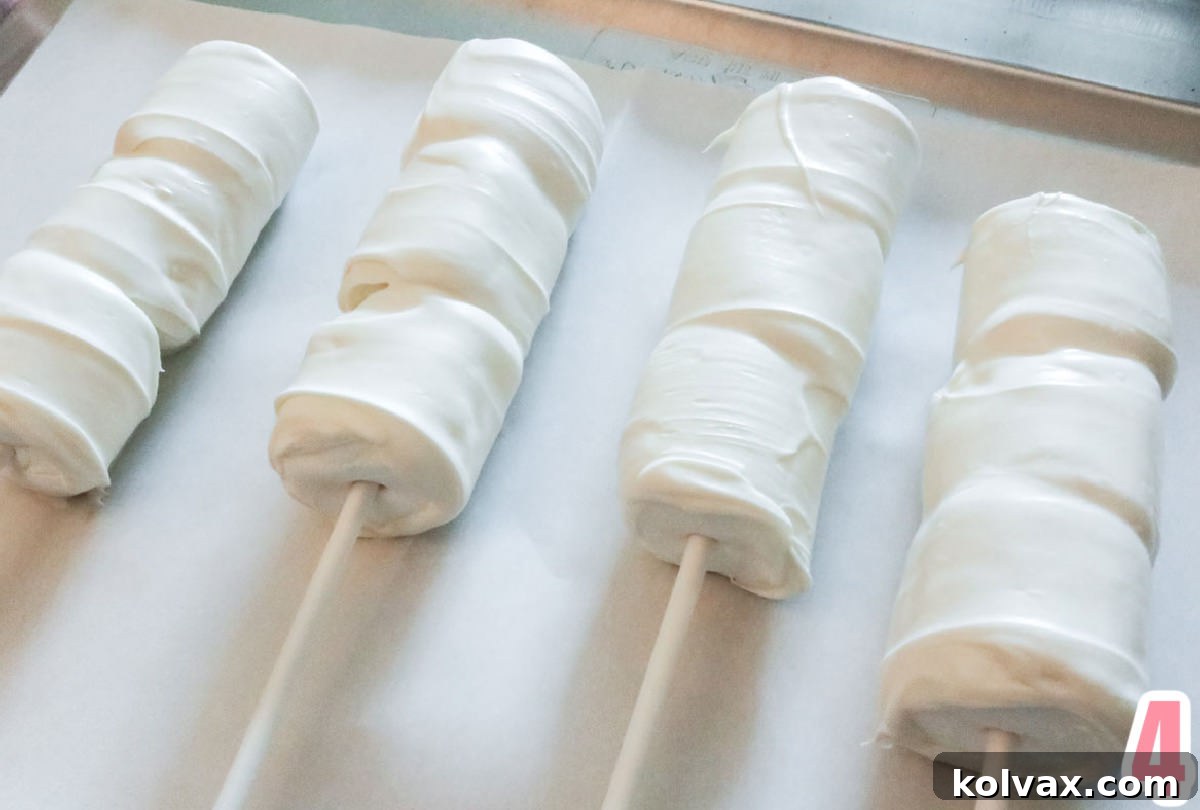 Four white chocolate-covered marshmallow pops drying on a parchment paper-lined cookie sheet, showing a smooth, even coating.