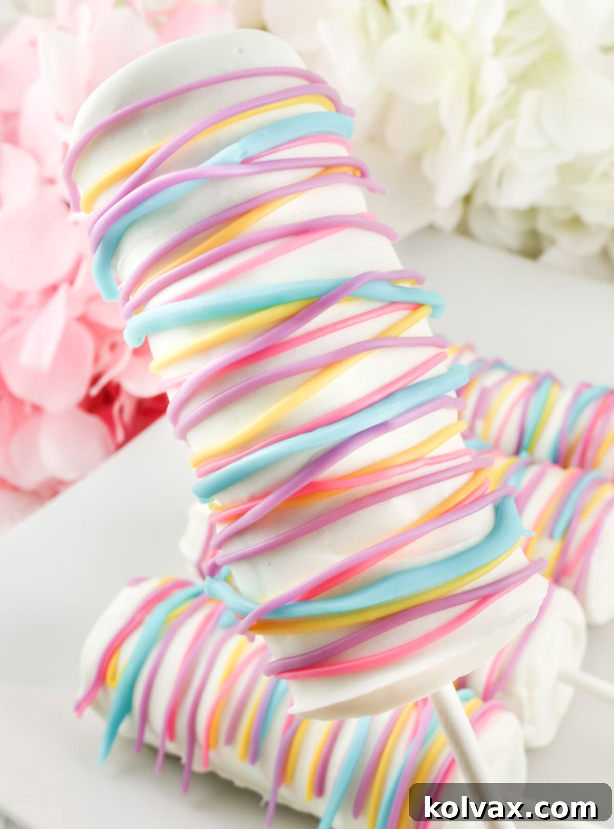 Close up of a White Chocolate Celebration Marshmallow Pop being held up by the lollipop stick over a plate of other pops, highlighting its smooth coating and vibrant drizzles.