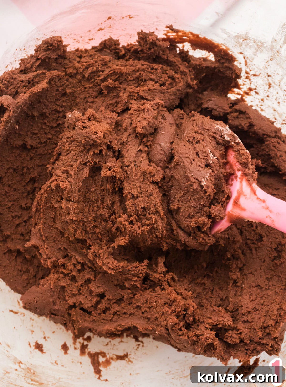 Closeup on a clear glass mixing bowl filled with rich chocolate cookie dough and a vibrant pink spatula. The dough looks smooth and ready for baking.
