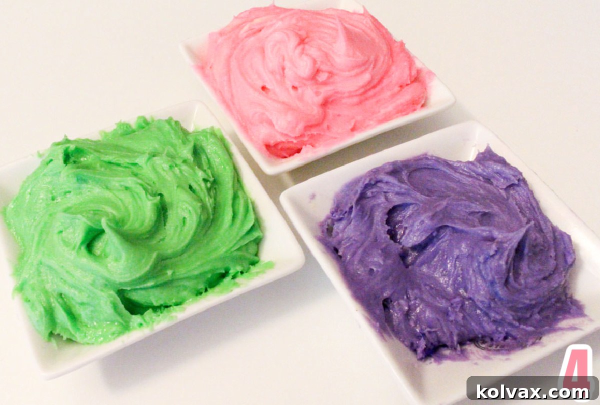 Three white ramekins filled with beautifully tinted buttercream frosting: one in soft pink, one in vibrant purple, and one in cheerful green. The frosting is smooth and ready for cookie decoration.