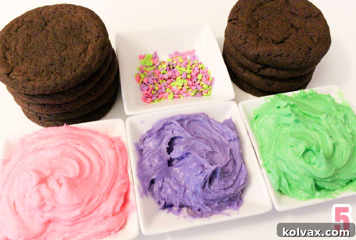 White ramekins filled with tinted pink, purple, and green buttercream frosting, alongside a bowl of colorful confetti sprinkles and two neat stacks of cooled chocolate cookies. All items are arranged on a white surface, ready for decorating.