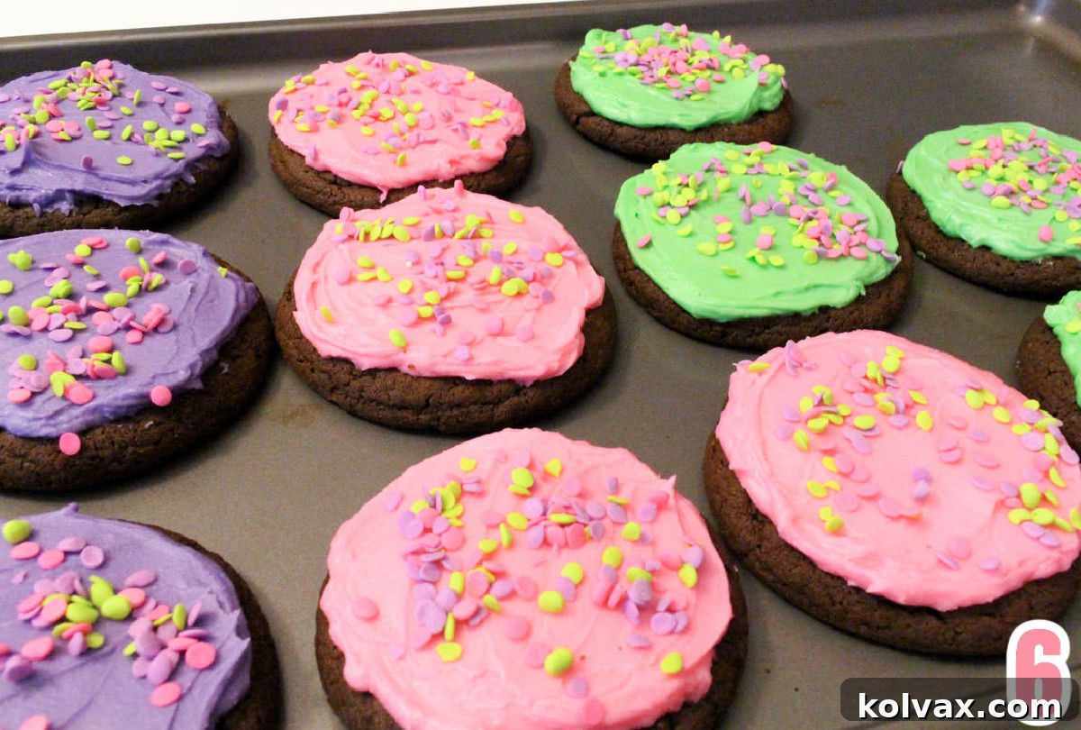 A cookie sheet filled with freshly frosted Frosted Easter Cookies. The chocolate cookies are generously covered in vibrant pink, purple, and green buttercream frosting, each adorned with a scattering of colorful confetti sprinkles, creating a cheerful display.