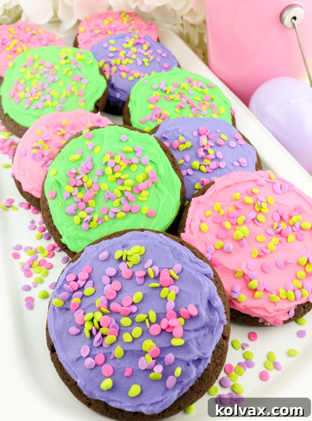 Closeup of a white platter abundantly filled with beautifully Frosted Easter Cookies. The cookies are decorated with pink, purple, and green buttercream and confetti sprinkles, artfully arranged alongside extra sprinkles and decorative plastic Easter eggs, creating a festive holiday display.