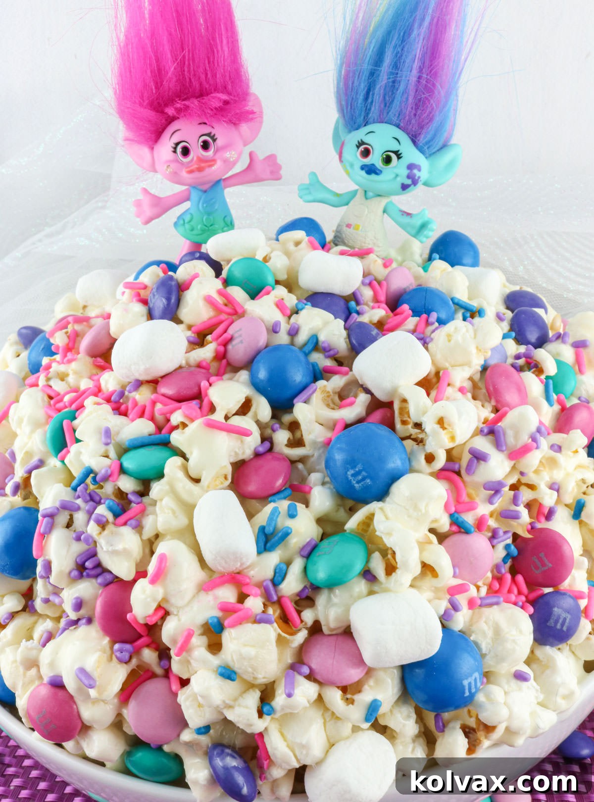 Closeup on a white serving bowl filled with Trolls Popcorn with two toy Trolls  sitting on the top of the bowl.