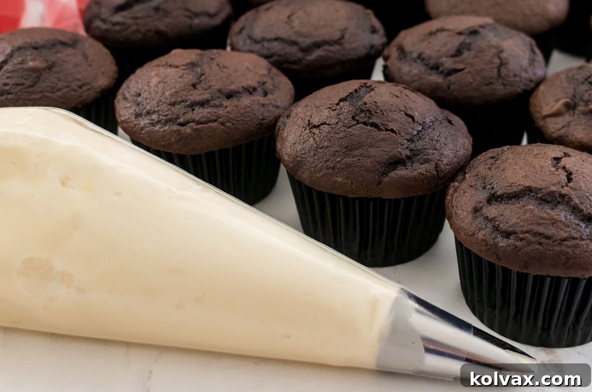 Decorating bag filled with Marshmellow Frosting sitting on a white table next to a batch of unfrosted chocolate cupcakes.