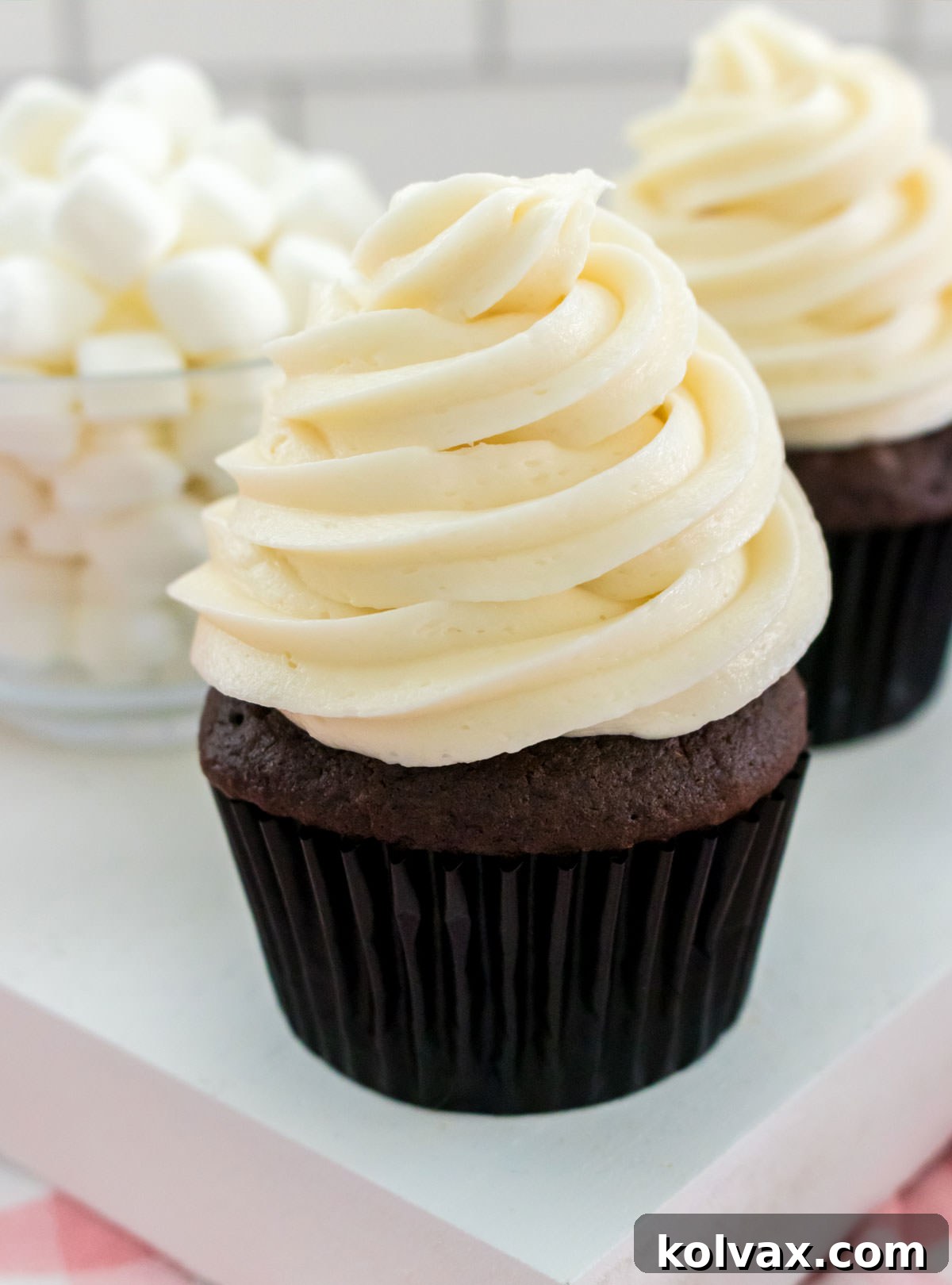 Closeup on two chocolate cupcakes topped with a large swirl of The Best Marshmallow Frosting sitting next to a glass bowl filled with mini marshmallows.