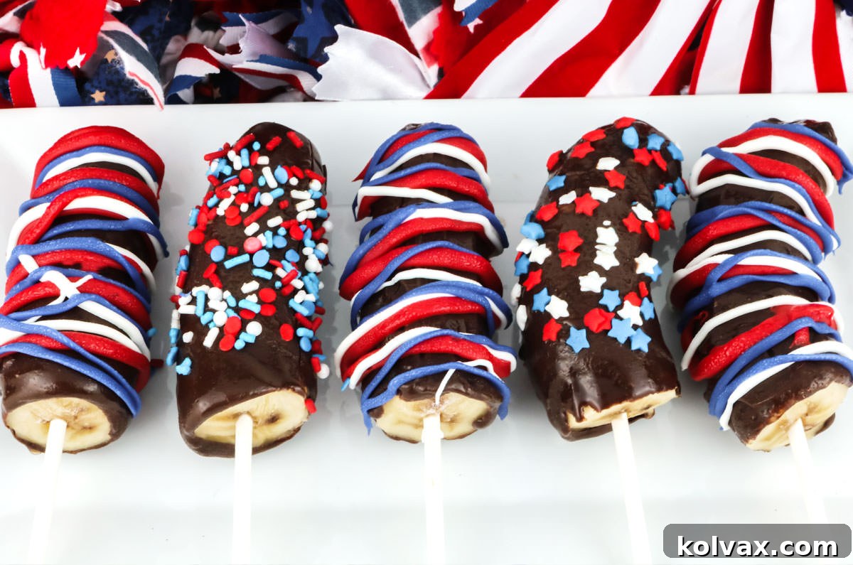 Five patriotic Frozen Chocolate-Covered Bananas decorated with red, white, and blue drizzles, presented on a white platter for a 4th of July party.