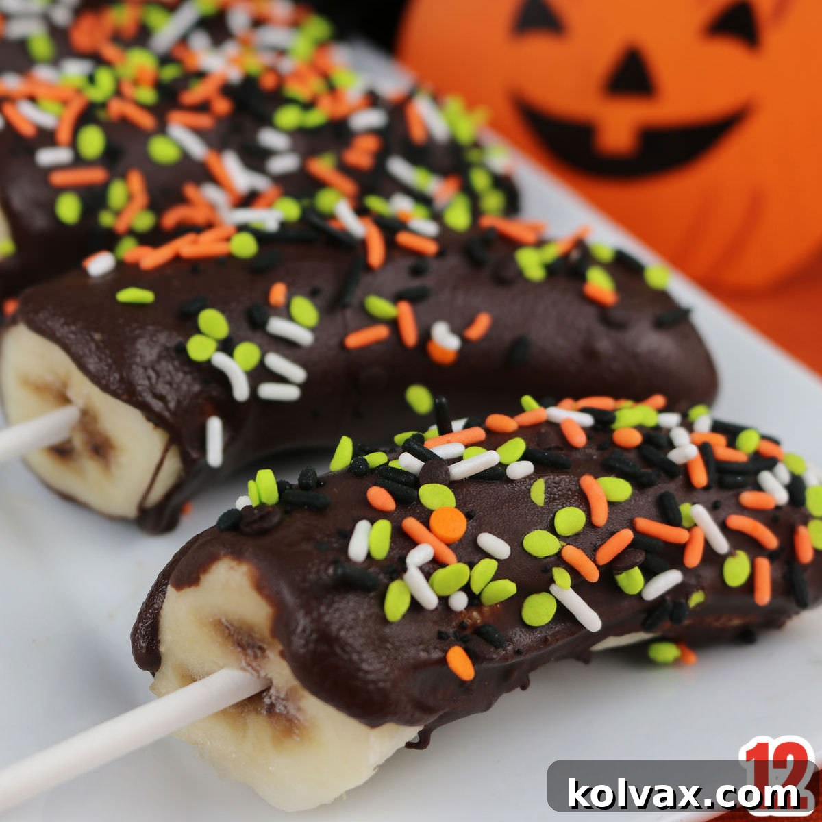 Close-up of four Halloween-themed Frozen Chocolate-Covered Bananas with orange drizzles and spooky sprinkles, placed on a white platter beside a carved pumpkin.