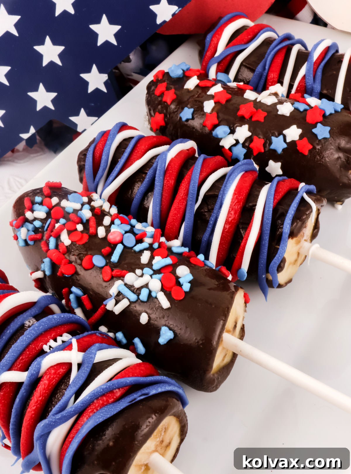 A beautiful arrangement of five patriotic Frozen Chocolate-Covered Bananas, decorated with red, white, and blue drizzles, presented on a white serving platter against 4th of July decorations.