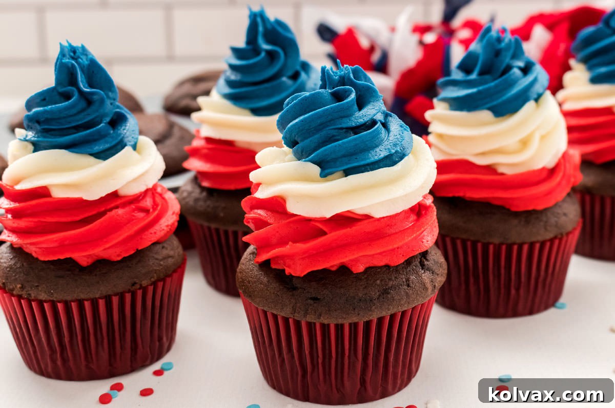 Five beautifully decorated Patriotic Swirl Cupcakes with vibrant red, white, and blue frosting, resting on a white table. In the background, a cupcake tin holds more chocolate cupcakes, suggesting the process of creation.