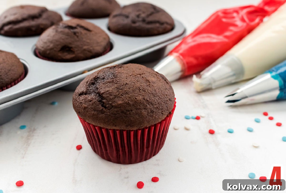 A single chocolate cupcake in a festive red liner stands on a white table, flanked by three individual decorating bags, each filled with red, white, and blue frosting. In the background, a cupcake tin is filled with additional cupcakes, awaiting their colorful adornment.