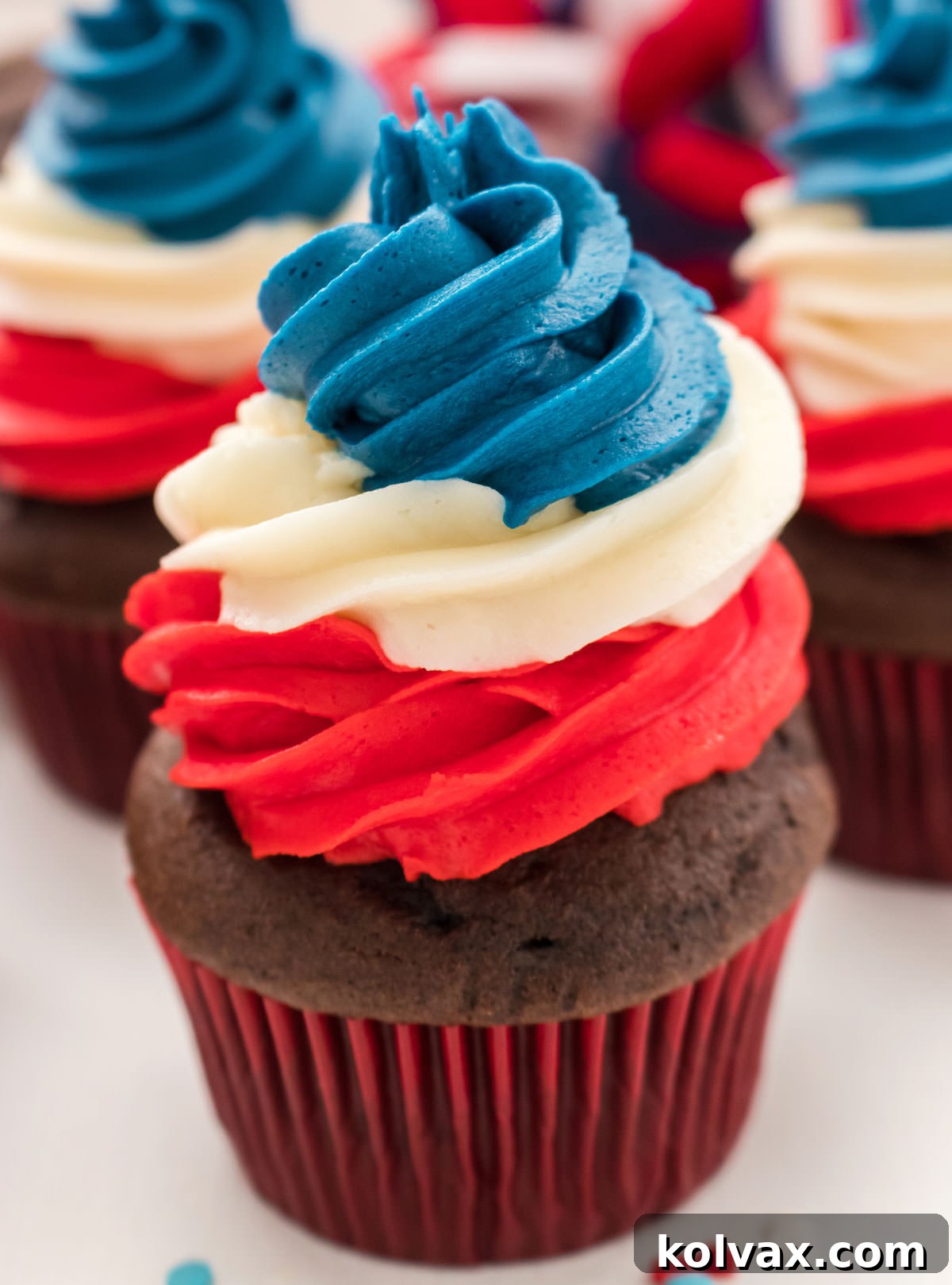 A stunning closeup of a single, perfectly frosted Patriotic Swirl Cupcake, adorned with luscious red, white, and blue buttercream frosting, prominently displayed in front of an array of other festive 4th of July cupcakes.