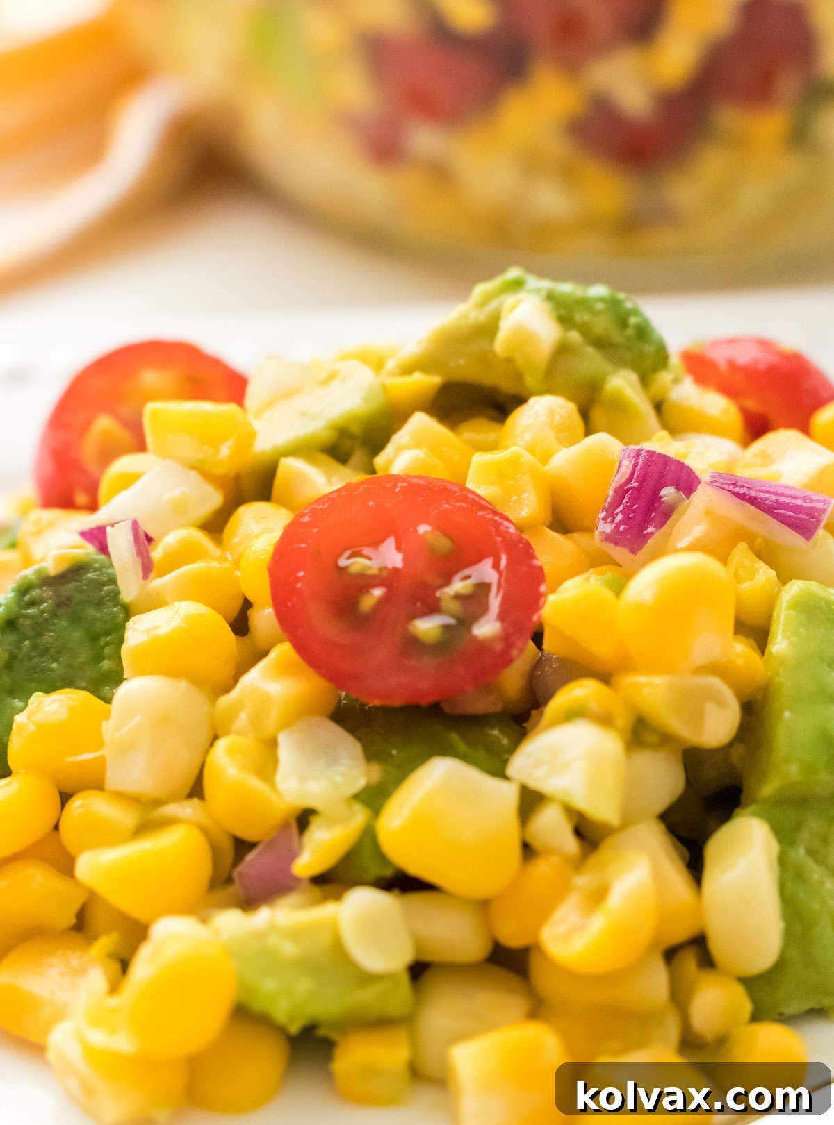 Closeup on a large helping of The Best Ever Summer Corn Salad sitting on a white plate on a white table in front of a glass bowl filled with Corn Salad.
