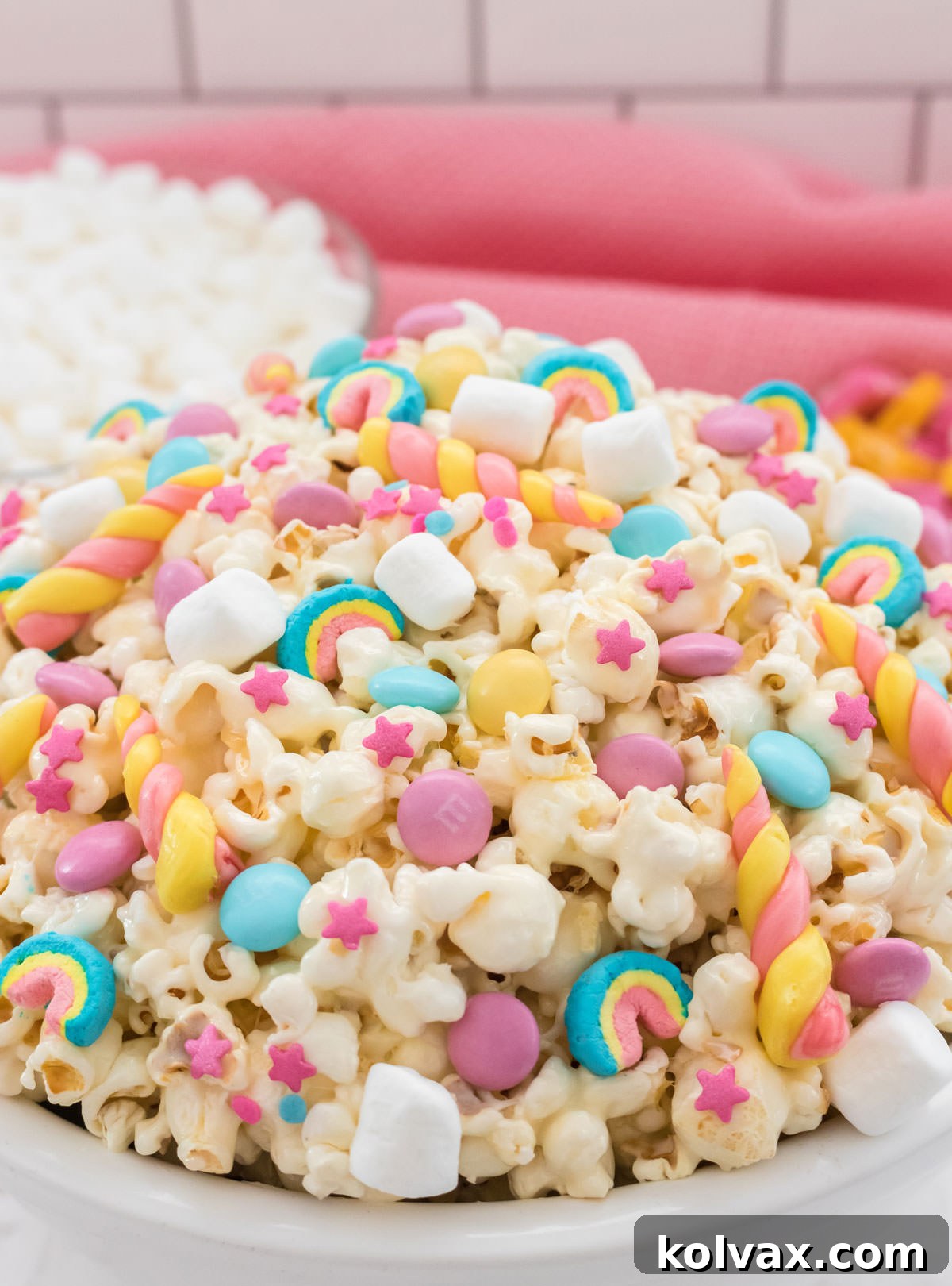 Closeup on a white serving bowl filled with colorful Unicorn Popcorn with a bowl of mini marshmallows in the background and a pink table linen.