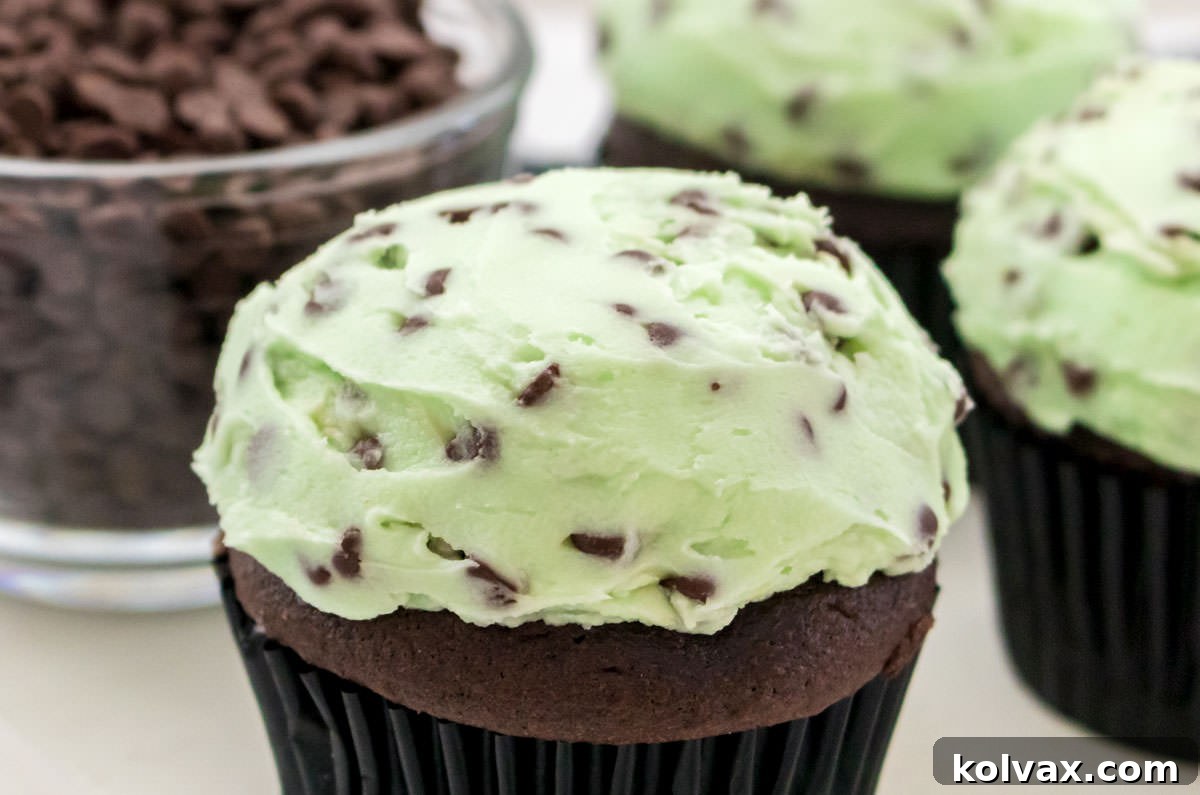 Closeup on cupcakes topped with vibrant Mint Chocolate Chip Buttercream Frosting, sitting on a pristine white surface beside a small ramekin brimming with mini chocolate chips.
