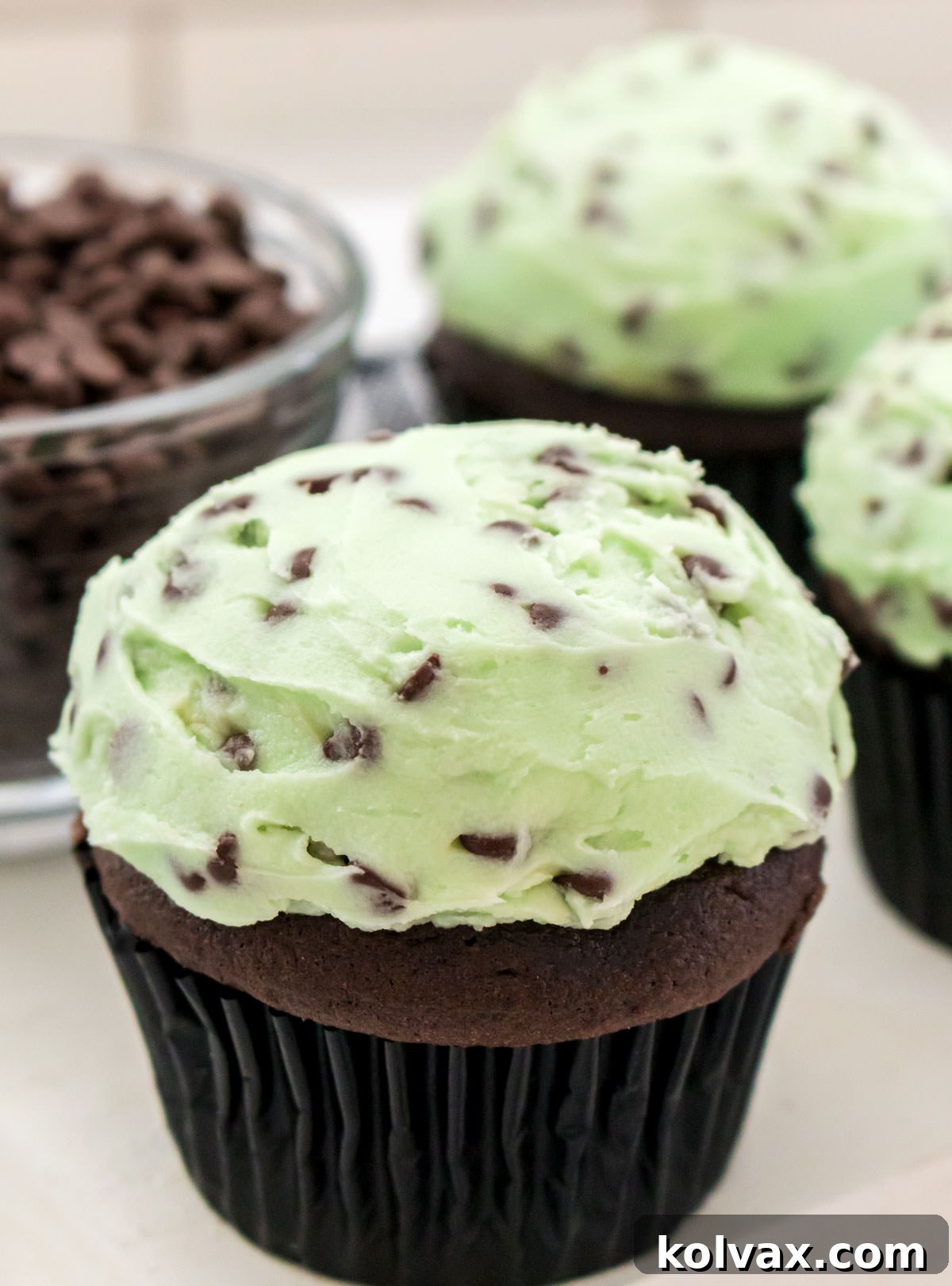 Closeup on three decadent chocolate cupcakes generously frosted with Mint Chocolate Chip buttercream, arranged beautifully on a white table beside a charming bowl overflowing with chocolate chips.