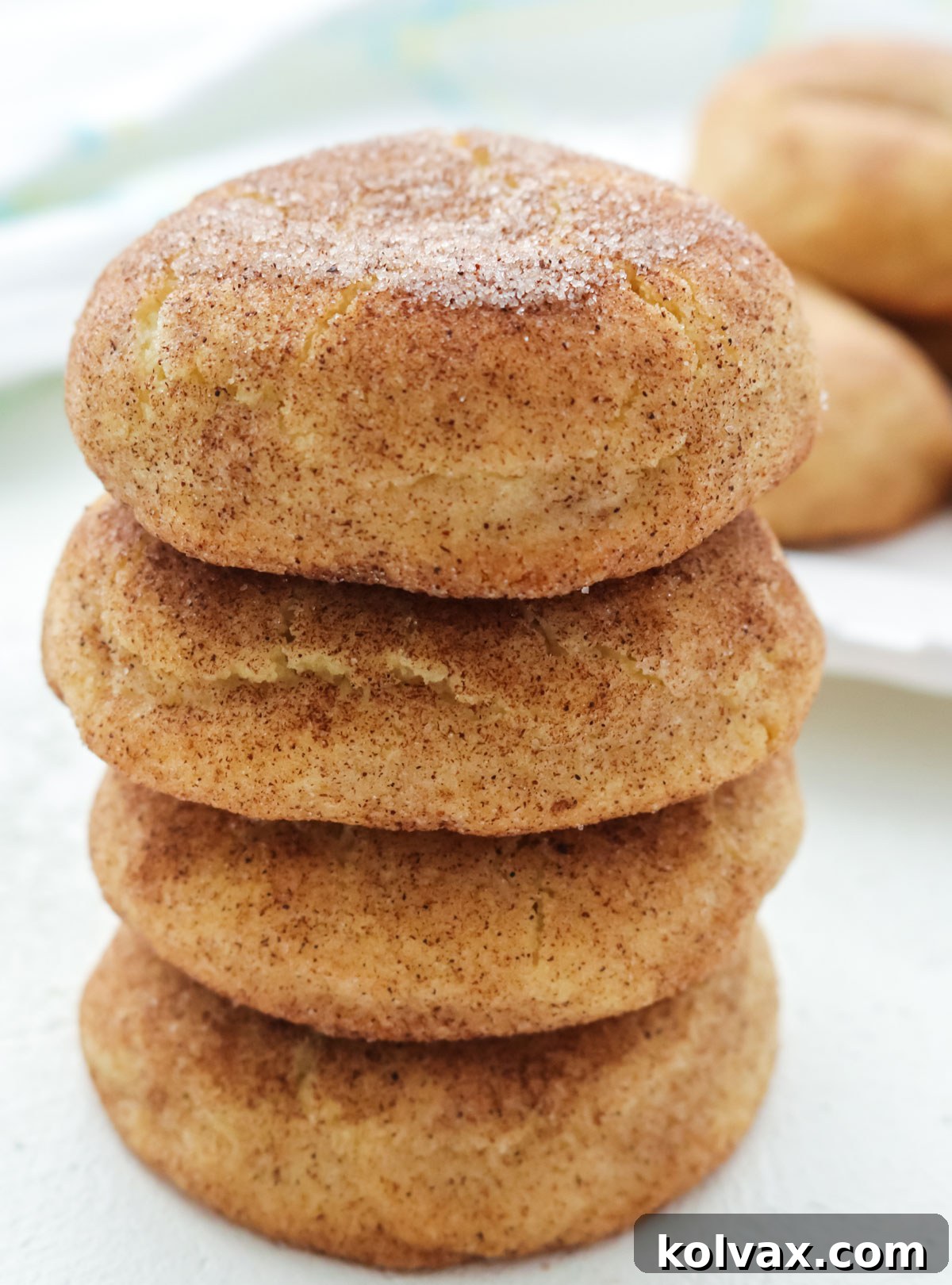 Melt-in-Your-Mouth Snickerdoodle Perfection 7 Closeup on a stack of four Snickerdoodle cookies sitting on a white table in front of a plate of Snickerdoodles.