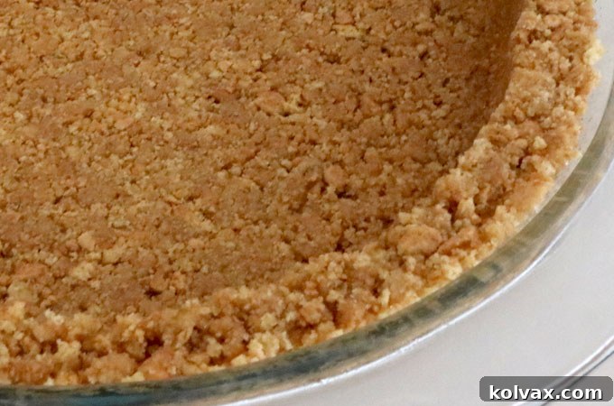 A hand using the flat bottom of a measuring cup to firmly press the moistened graham cracker crumbs into the bottom and up the sides of a pie plate, creating a solid crust.