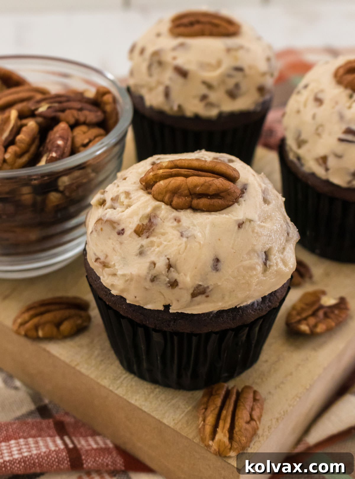 Closeup on three chocolate cupcakes topped with Maple Pecan Buttercream Frosting sitting on a wooden cutting board next to a glass bowl filled with pecans.