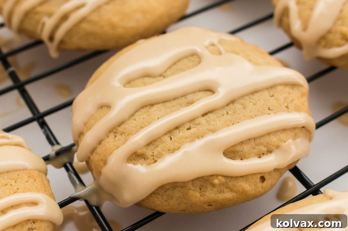 Closeup on a perfectly baked Maple Cookie, generously coated with creamy maple icing, resting on a cooling rack over a pristine white surface. The golden brown edges and the smooth, glistening glaze highlight its delicious appeal.
