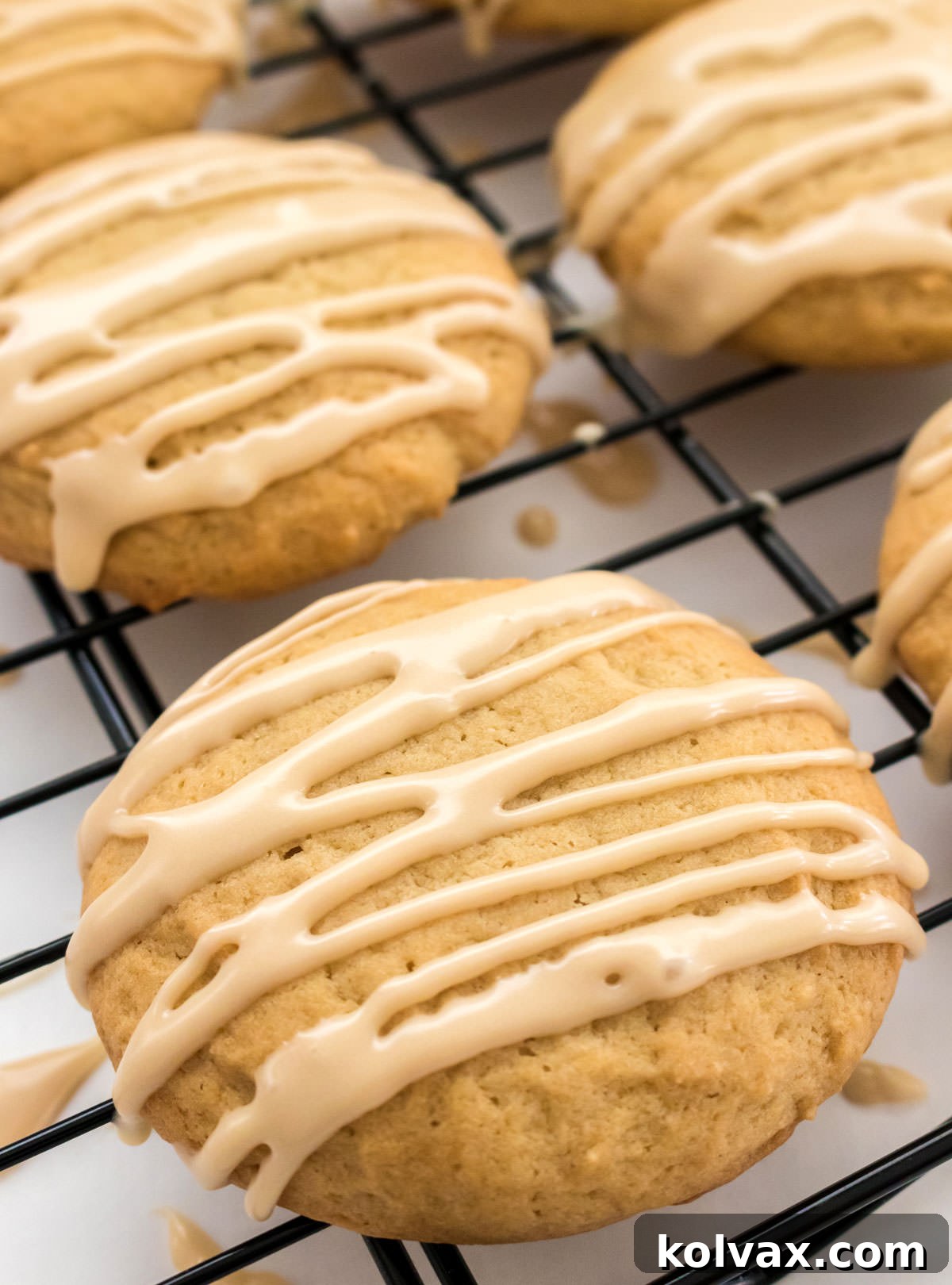 A close-up shot of a fresh batch of golden Maple Cookies, generously coated with creamy maple frosting, neatly arranged on a black cooling rack. The glistening glaze and soft cookie texture create an inviting and delectable display.