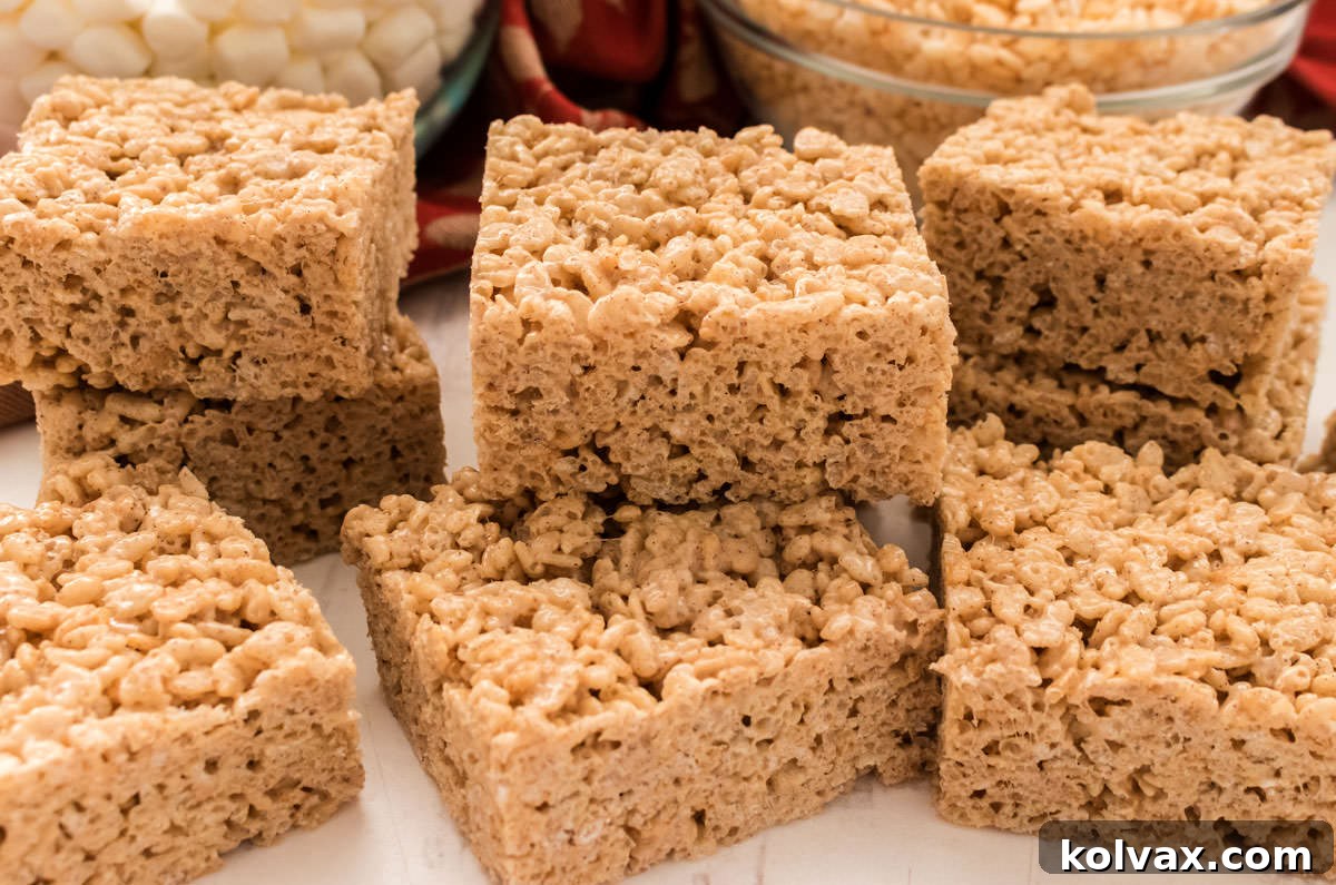 Closeup on ten Pumpkin Spice Rice Krispie Treats sitting on a white table in front of bowls of mini marshmallows and Rice Krispie Cereal.