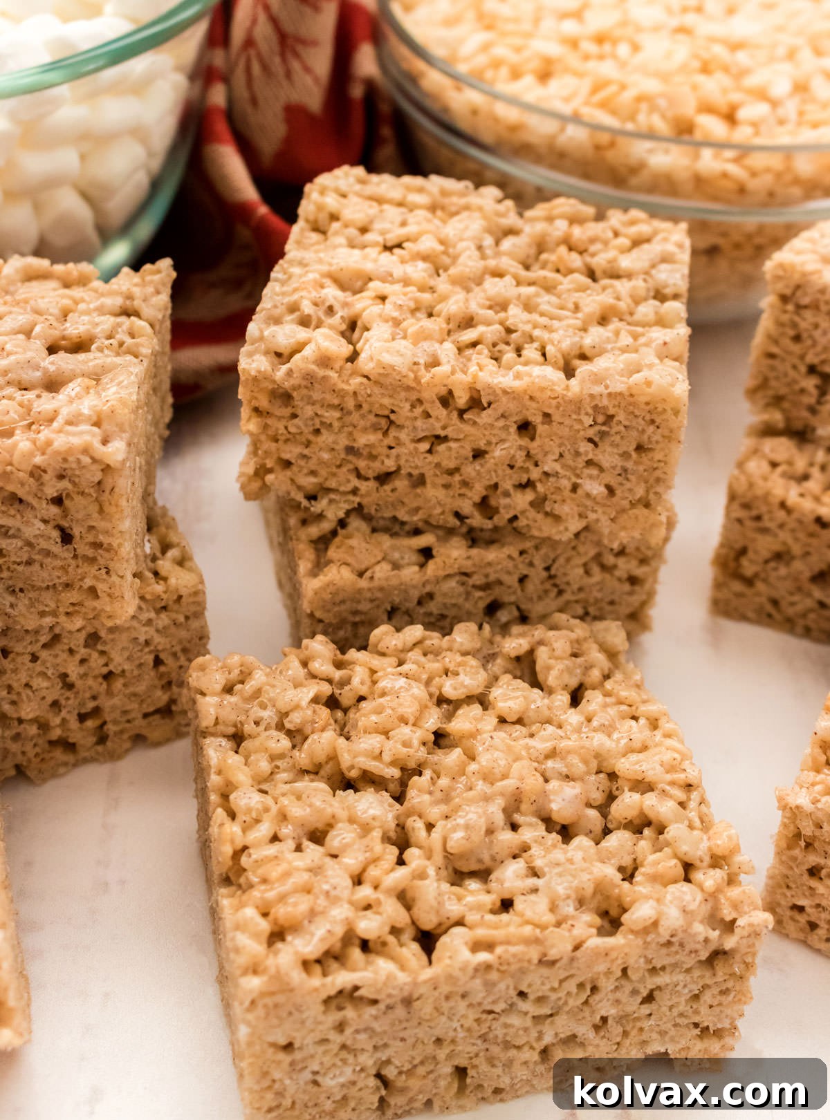 Stacks of Pumpkin Spice Rice Krispie Treats sitting on a white surface in front of bowls of mini marshmallows and Rice Krispie Cereal.