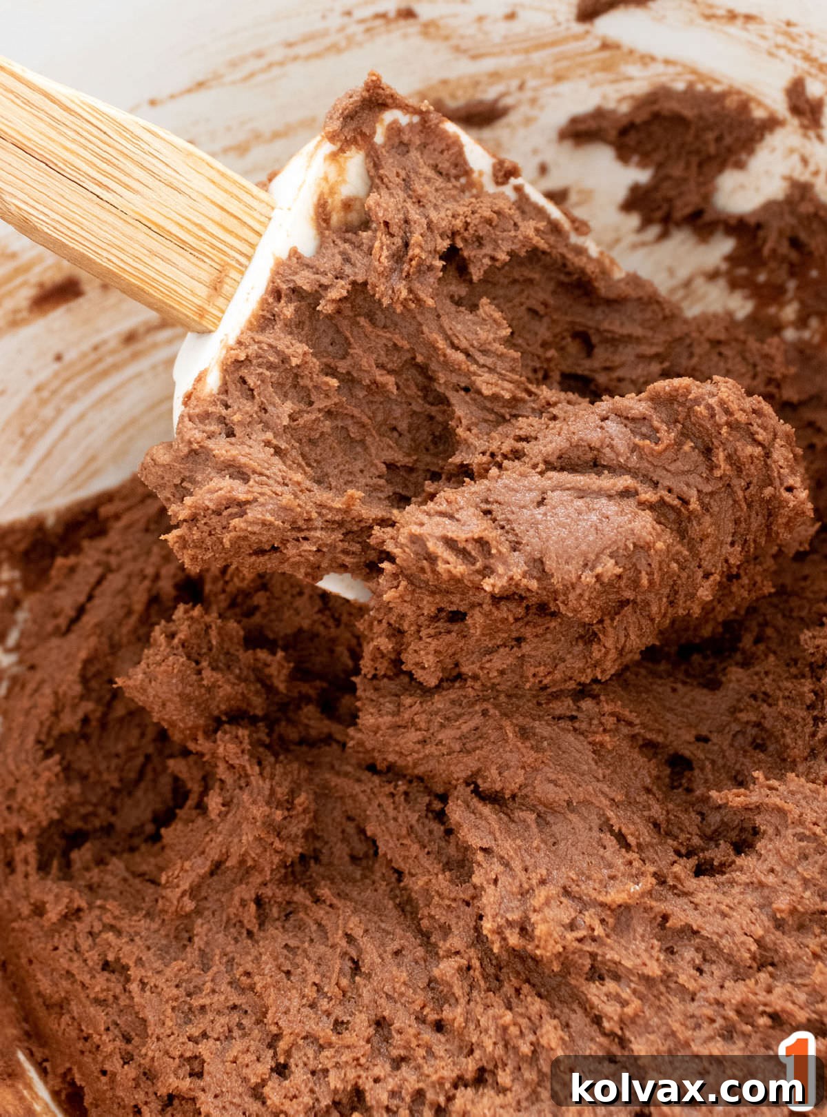 Closeup on a white mixing bowl filled with rich chocolate cookie dough and a white wooden spatula, ready for baking.