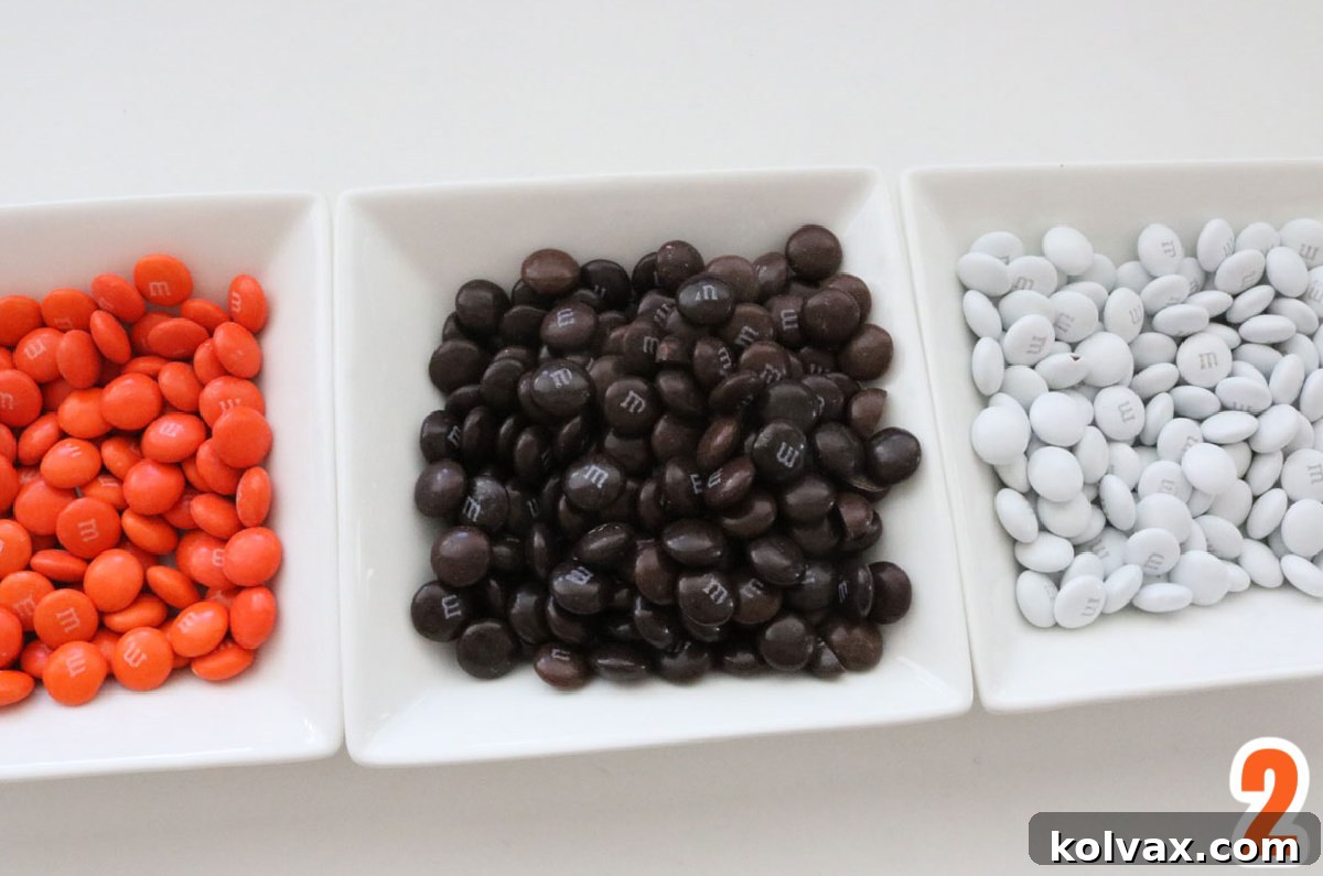 Three small white ramekins, each filled with a different color of M&M's: vibrant orange, rich dark brown, and crisp white.