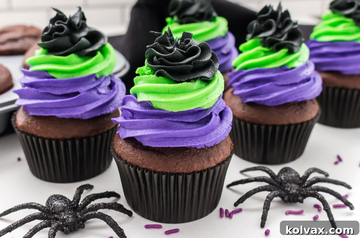 Closeup on five Bewitched Halloween Cupcakes sitting on a white table surrounded by toy spiders, black towel and a cupcake pan filled with chocolate cupcakes.