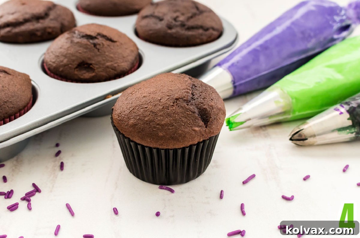 Chocolate Cupcake sitting on a white table surrounded by a cupcake pan filled with cupcakes and decorating bags filled with purple, green and black frosting.