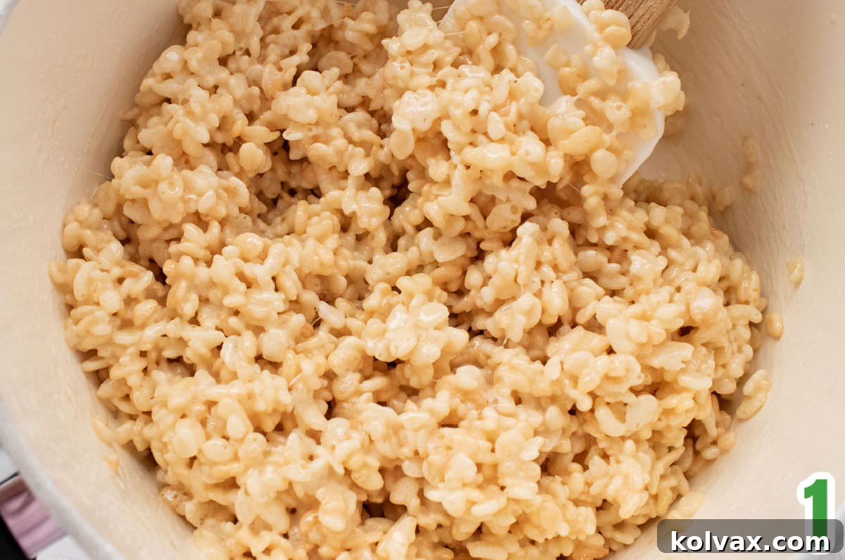 Closeup on a white saucepan filled with warm, gooey Rice Krispie Treat mixture and a white wooden spatula, ready for coloring.