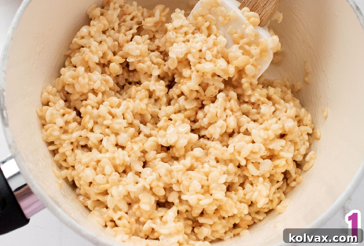 Close-up view of a white saucepan filled with the golden, gooey Rice Krispie Treat mixture, with a white wooden spoon resting inside.
