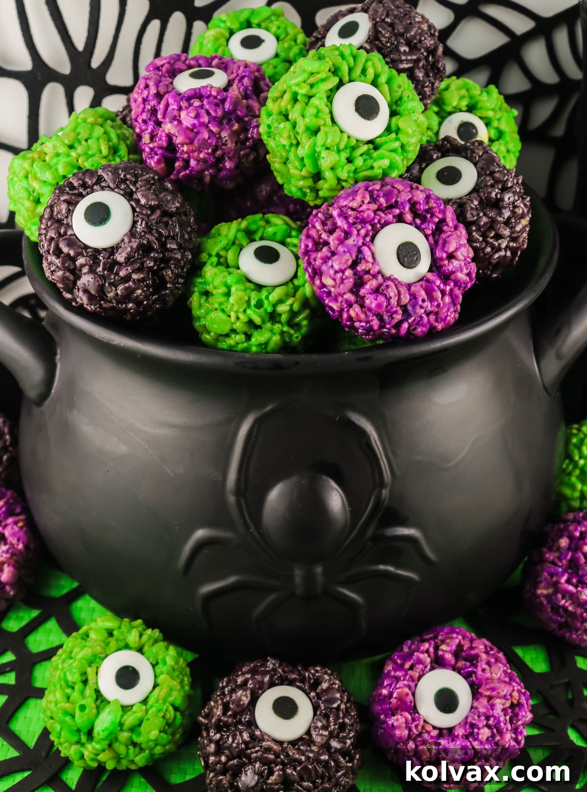 Close-up on a decorative black witches cauldron overflowing with an assortment of colorful Monster Eye Balls Rice Krispie Bites in green, purple, and black hues.